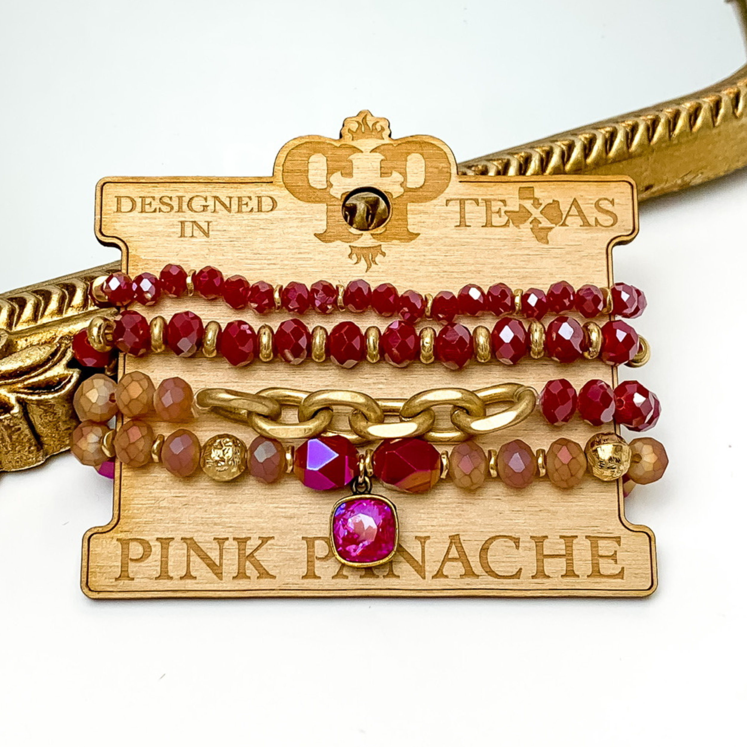 Set of four crystal beaded bracelets. The top two bracelets have gold beaded spacers. The middle bracelet has a gold chain segment. The last bracelet has a handing royal red delight cushion cut crystal. These bracelets are pictured on a wooden bracelet holder on a white background with a gold mirror behind them. 