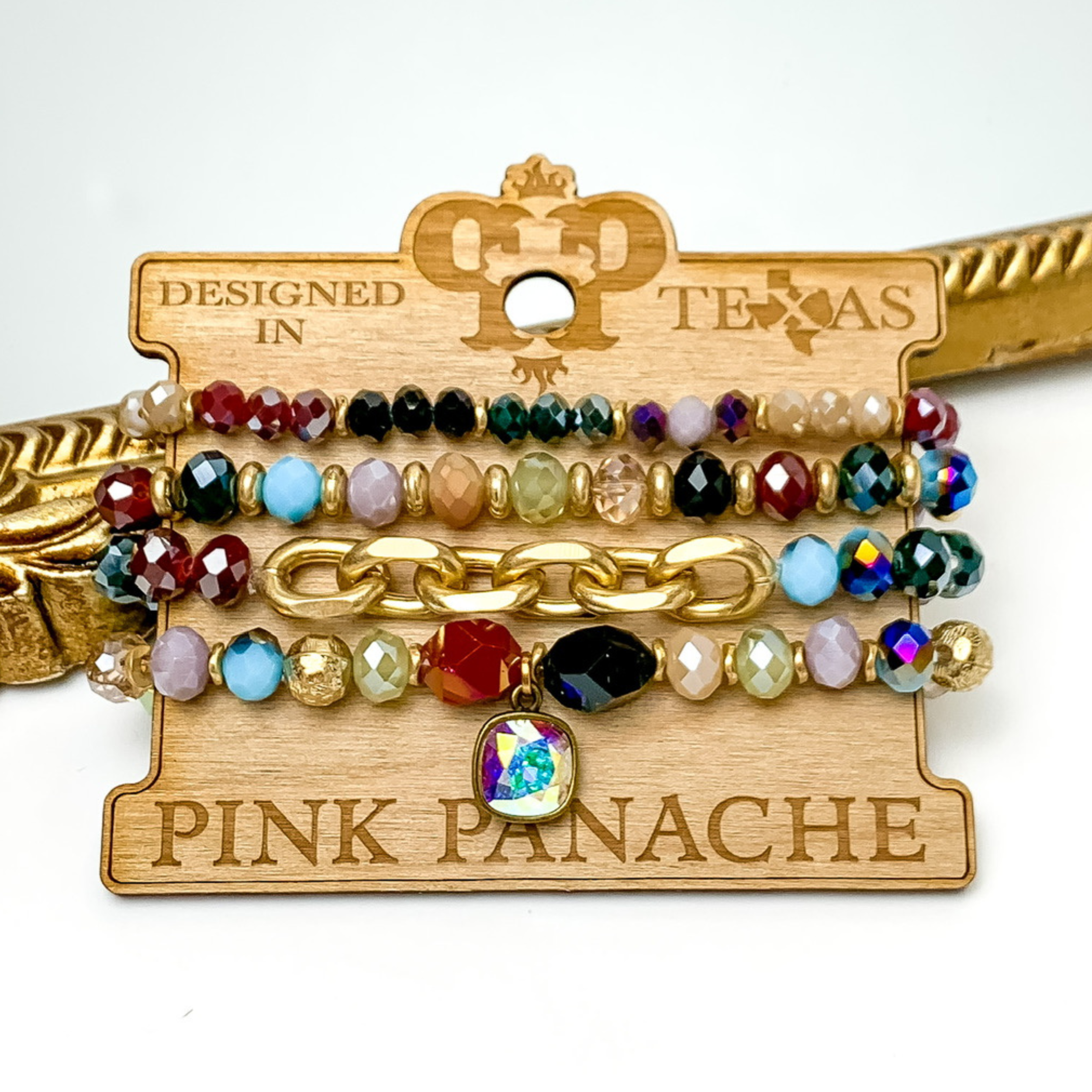 Set of four multicolored crystal beaded bracelets. The top two bracelets have gold beaded spacers. The middle bracelet has a gold chain segment. The last bracelet has a handing ab cushion cut crystal. These bracelets are pictured on a wooden bracelet holder on a white background with a gold mirror behind them. 