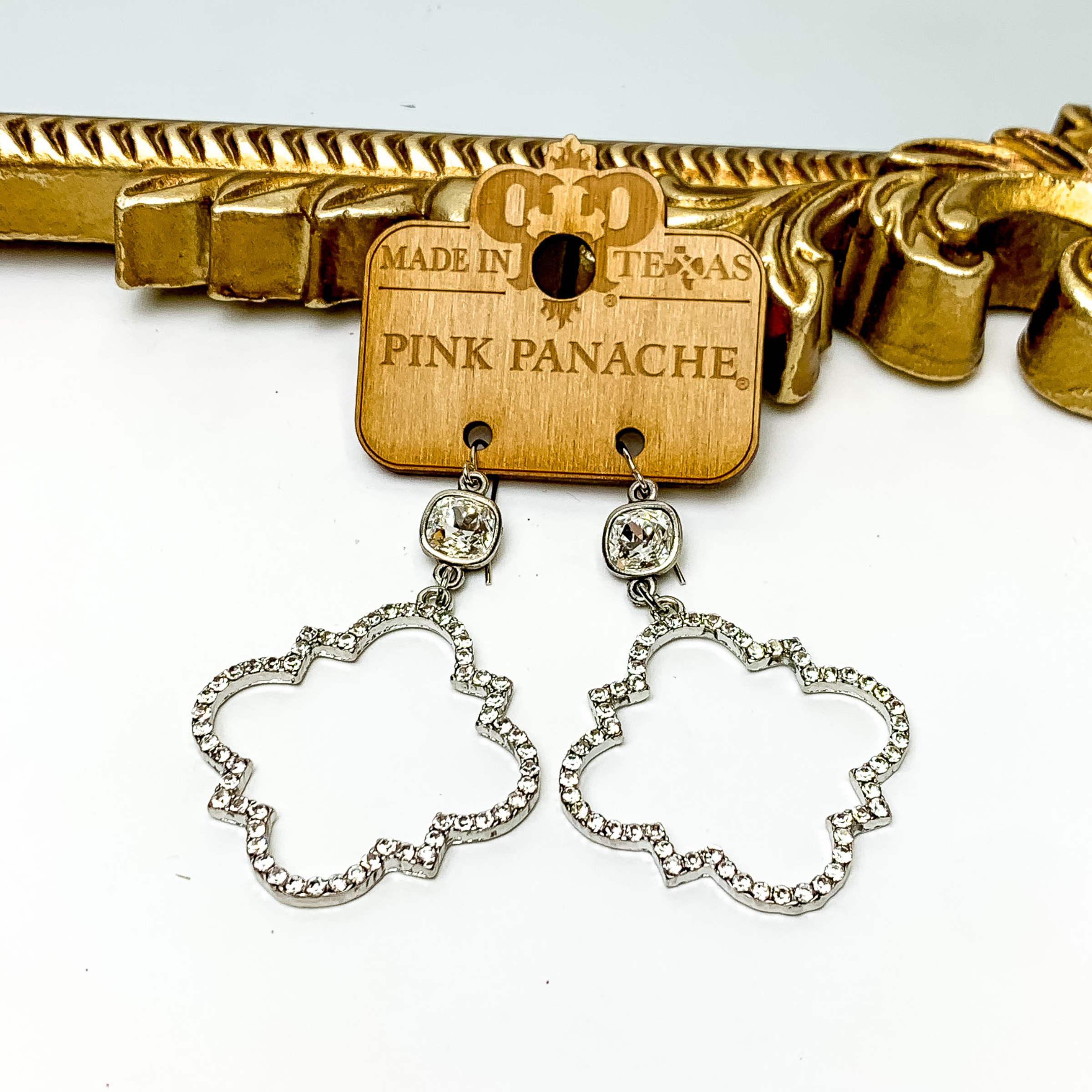 Clear cushion cut crystal drop earrings with a quatrefoil drop in silver. The quatrefoil has a clear crystal inlay. These earrings are pictured on a Pink Panache wood holder in front of a gold mirror and on a white background. 