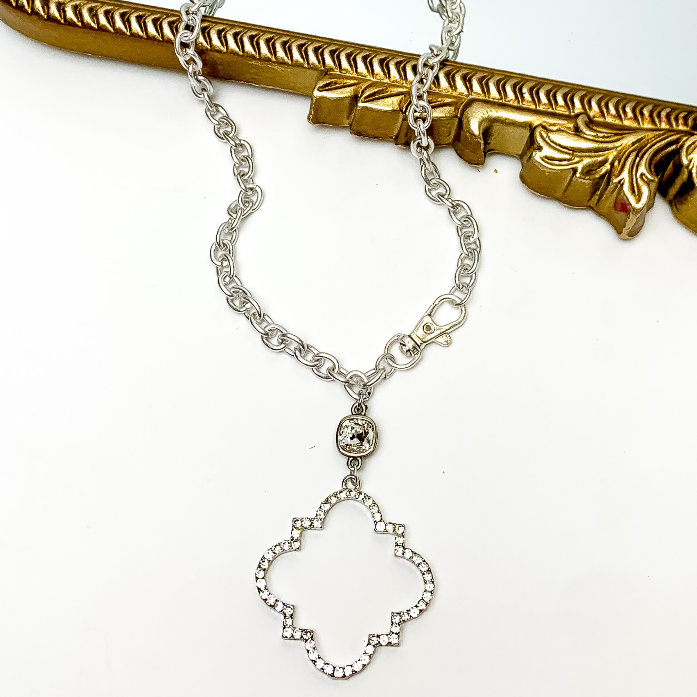 Chain necklace with a clear cushion cut crystal and quatrefoil drop in silver. The quatrefoil has a clear crystal inlay. This necklace is pictured patially laying on a gold mirror and on a white background. 