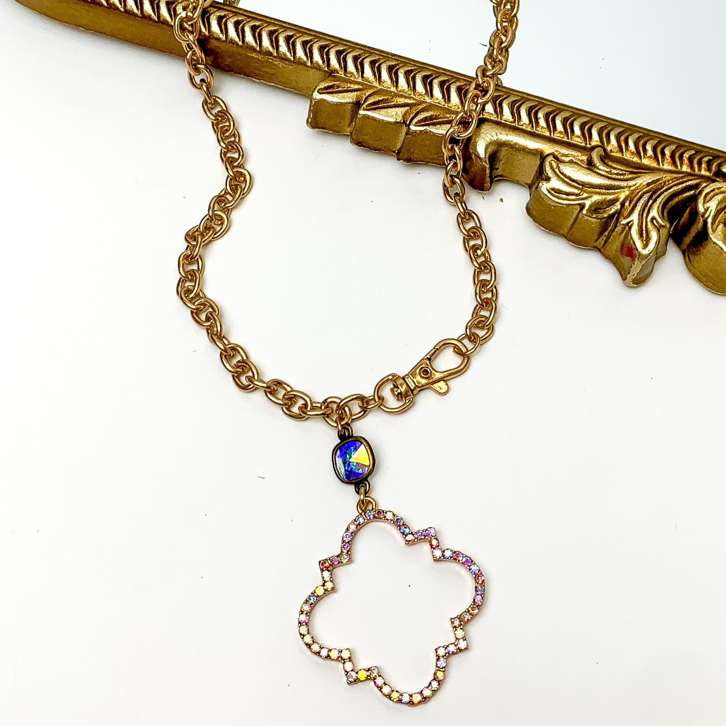 Chain necklace with a ab cushion cut crystal and quatrefoil drop in gold. The quatrefoil has a ab crystal inlay. This necklace is pictured patially laying on a gold mirror and on a white background. 