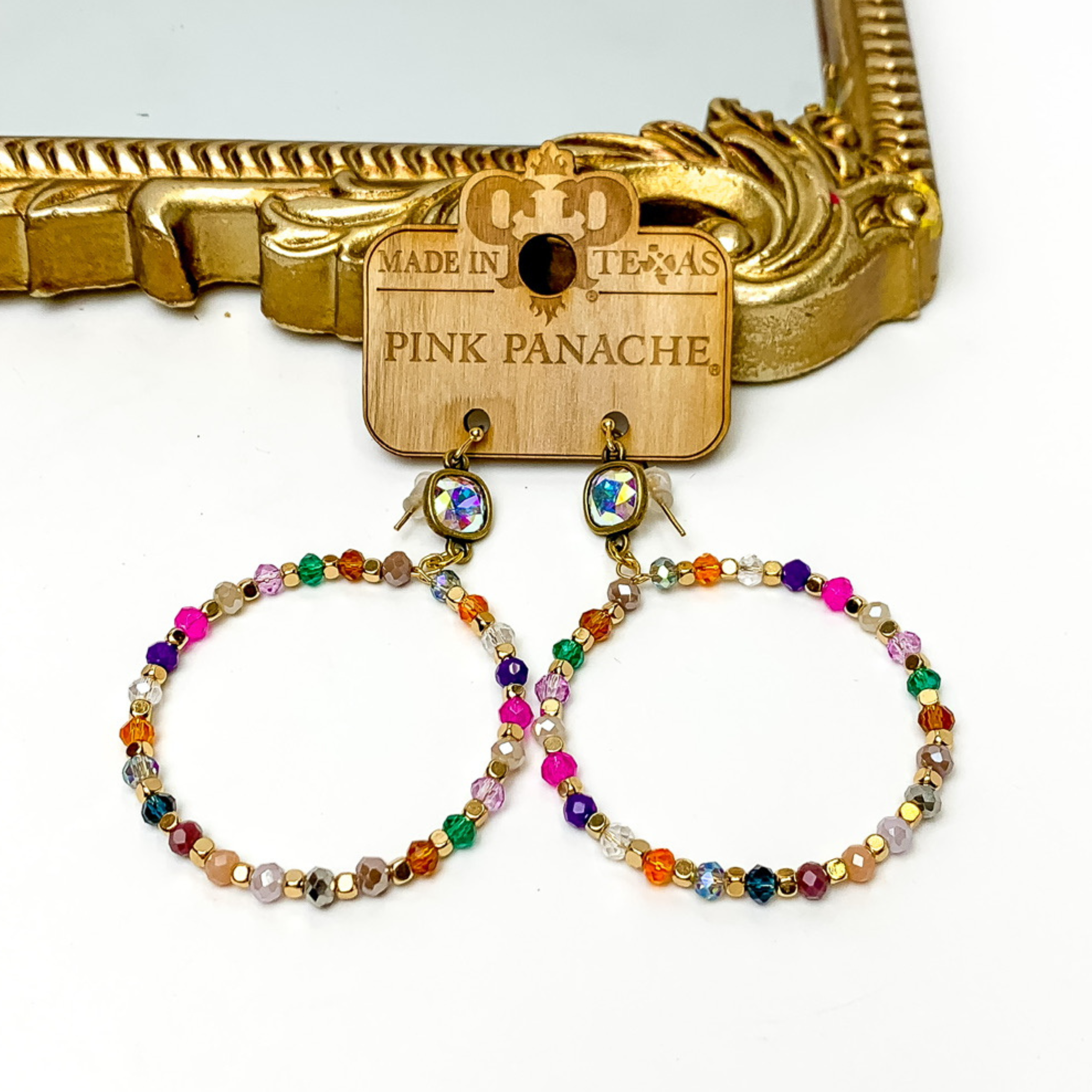 AB cushion cut crystal drop earrings with a multicolored, beaded circle pendant. These earrings are pictured on a Pink Panache wood holder in front of a gold mirror and on a white background. 