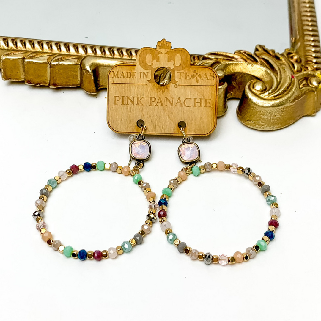 Rose water opal cushion cut crystal drop earrings with a multicolored, beaded circle pendant. These earrings are pictured on a Pink Panache wood holder in front of a gold mirror and on a white background. 