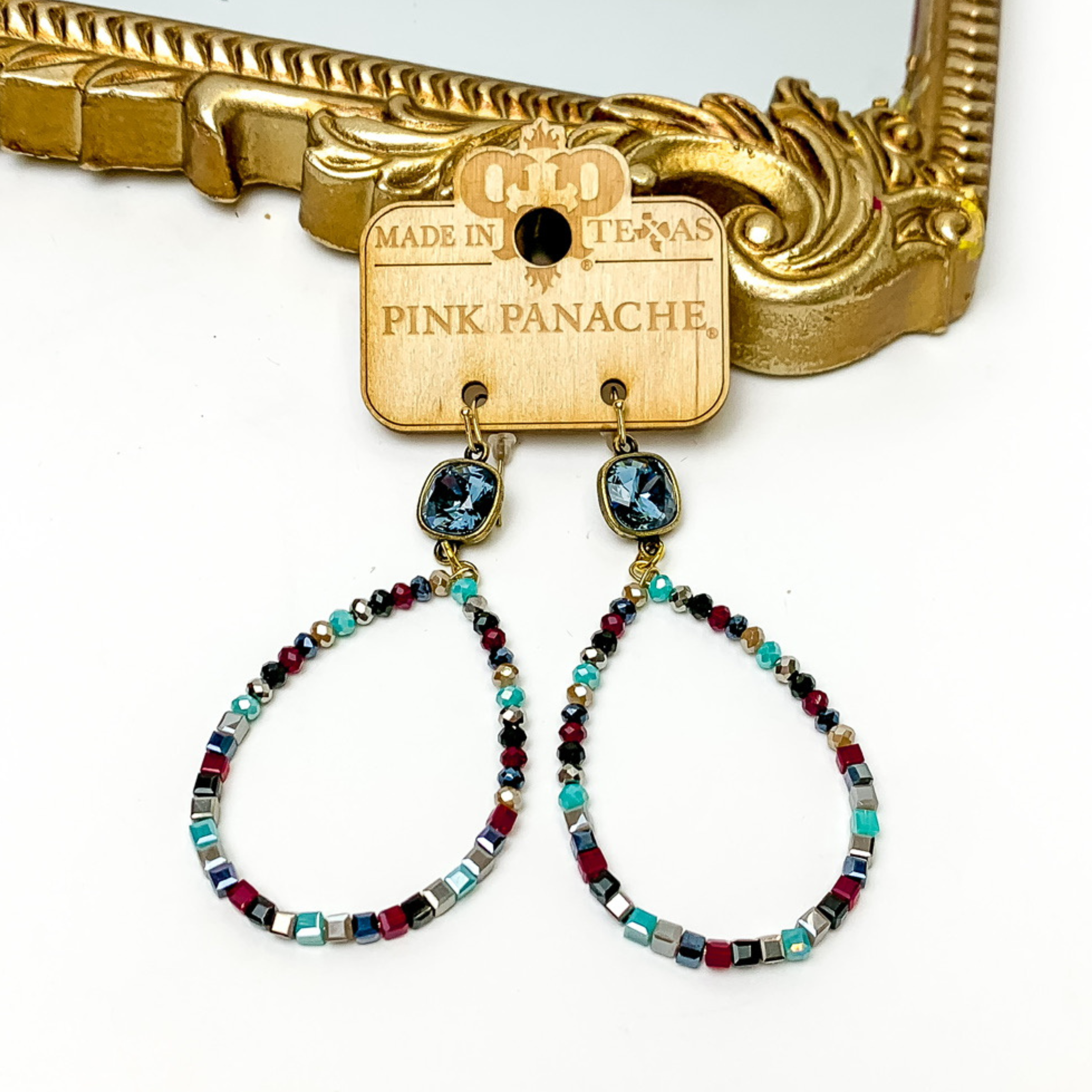 Denim blue cushion cut crystal drop earrings with a multicolored, beaded teardrop pendant. These earrings are pictured on a Pink Panache wood holder in front of a gold mirror and on a white background. 