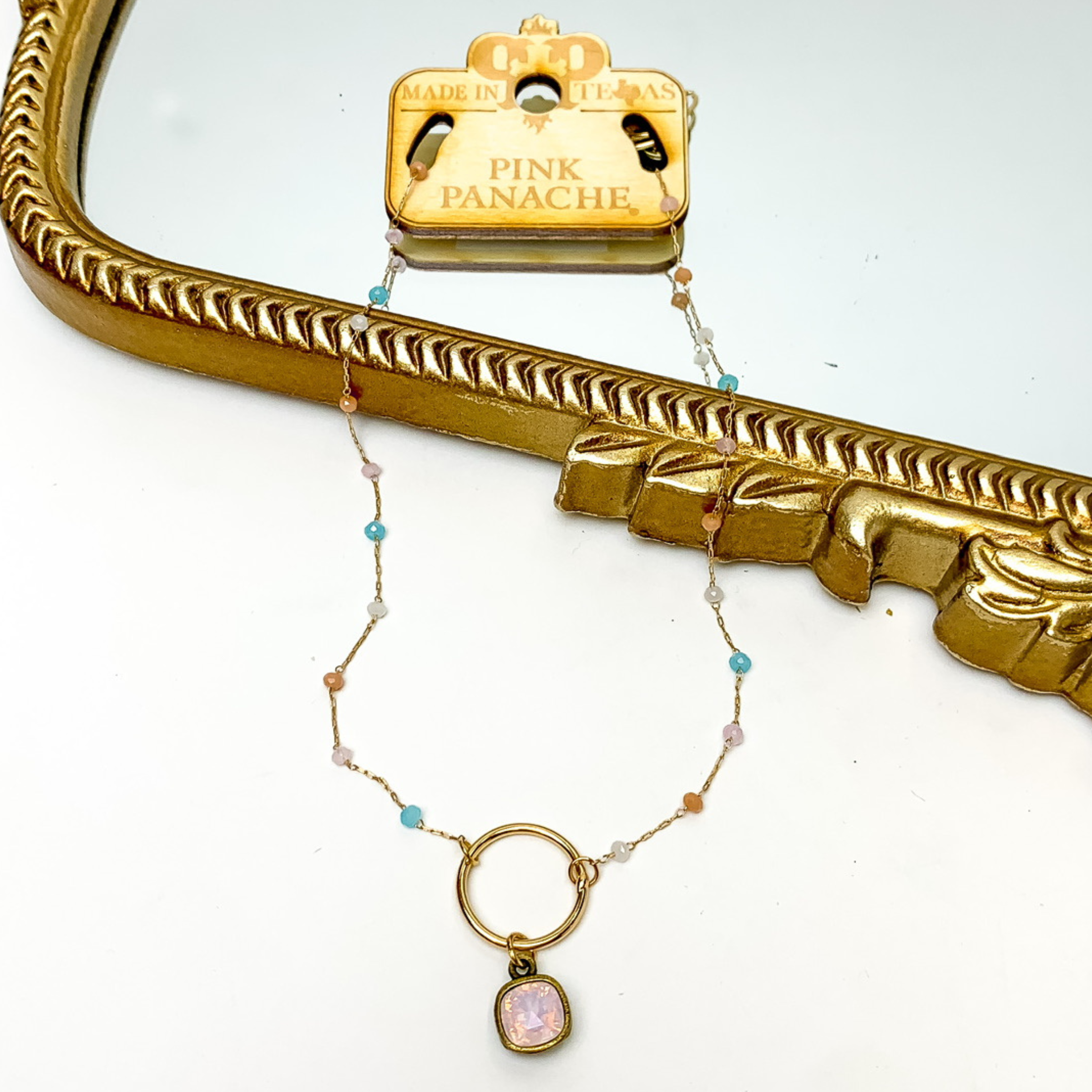 Gold chain necklace with multicolor, crystal beaded spacers. This necklace also includes a gold, open circle pendant with a rose water opal cushion cut crystal. This necklace is pictured on a wooden necklace tag and patially laying on a gold mirror and on a white background. 