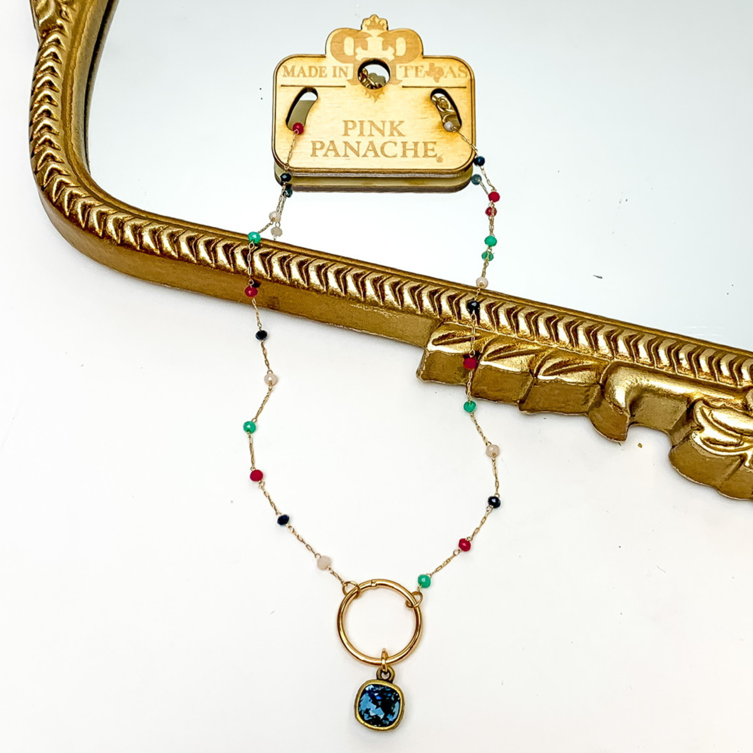 Gold chain necklace with multicolor, crystal beaded spacers. This necklace also includes a gold, open circle pendant with a denim blue cushion cut crystal. This necklace is pictured on a wooden necklace tag and patially laying on a gold mirror and on a white background. 