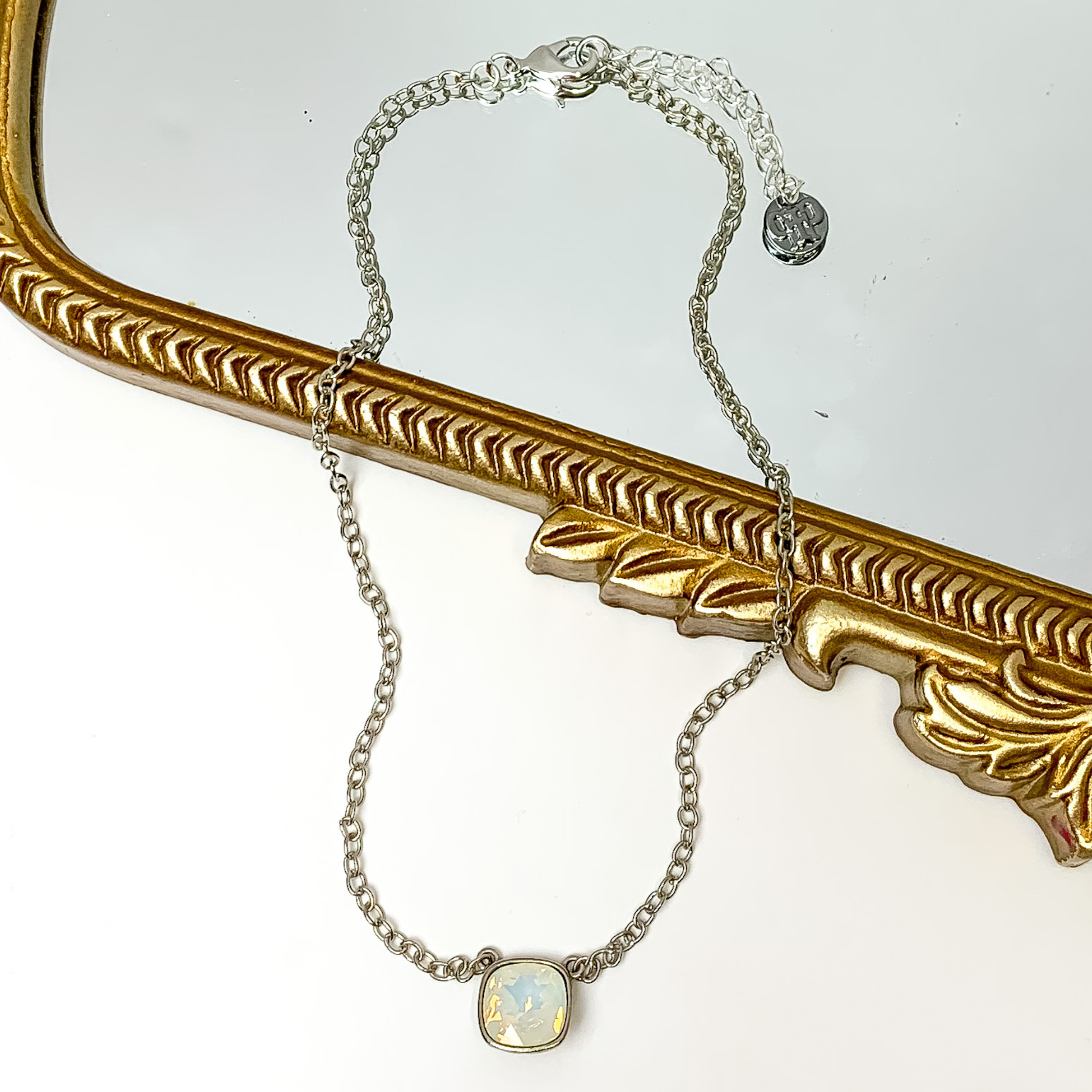 Silver chain necklace with a white opal cushion cut crystal. This necklace is pictured partially laying on a gold mirror on a white background.