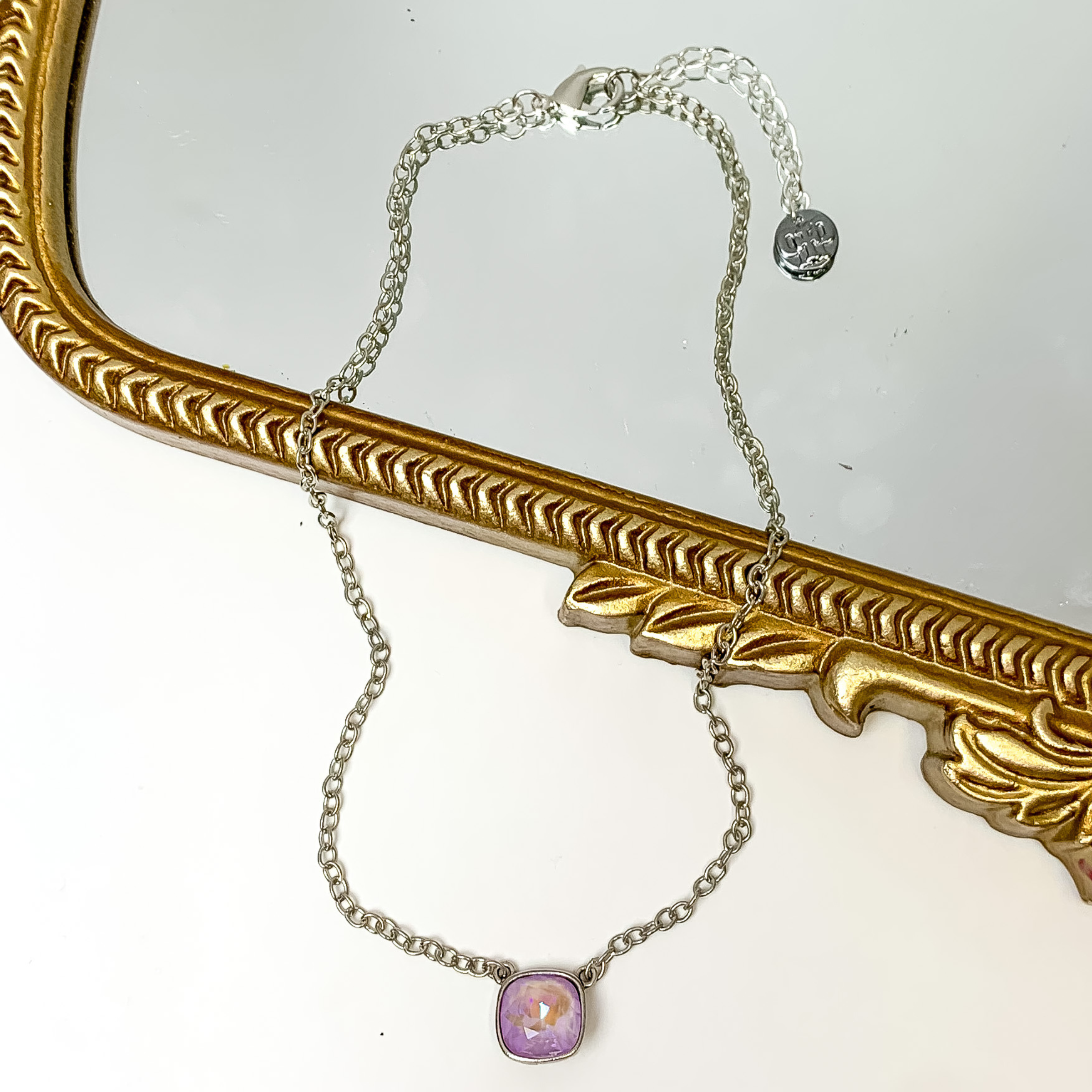 Silver chain necklace with a lavender cushion cut crystal. This necklace is pictured partially laying on a gold mirror on a white background.