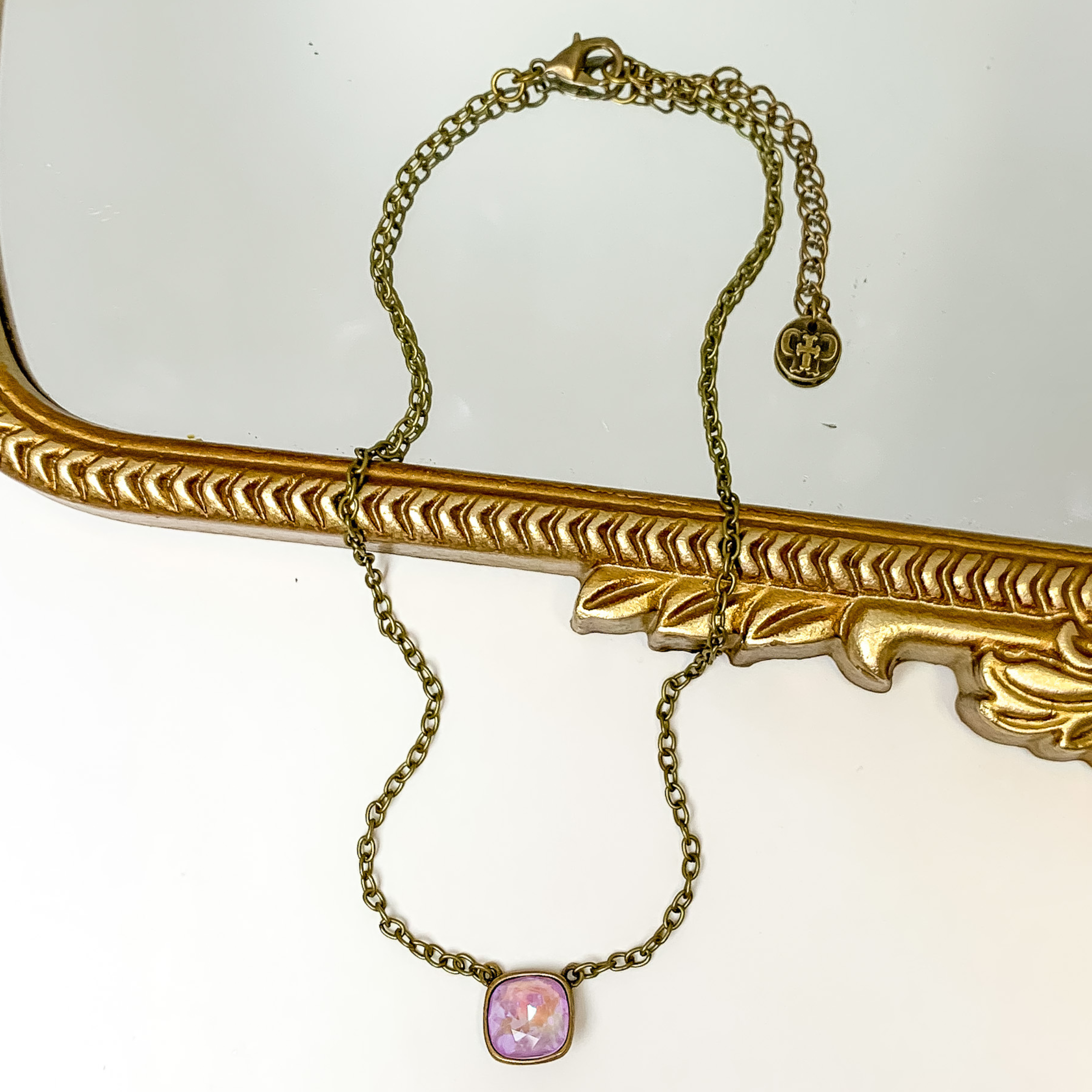 Bronze chain necklace with a lavender cushion cut crystal. This necklace is pictured partially laying on a gold mirror on a white background.