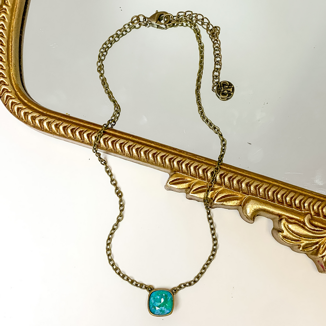 Bronze chain necklace with a laguna delight cushion cut crystal. This necklace is pictured partially laying on a gold mirror on a white background.