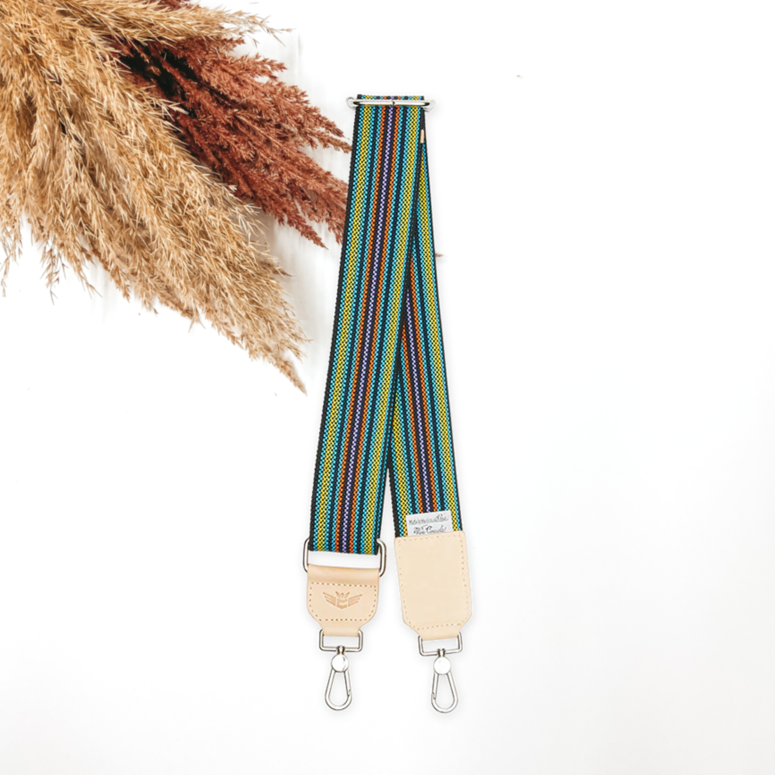 A woven multicolored striped purse strap that has a main color of black. This purse strap is pictured on a white background with tan and brown pompous grass in the top left corner.