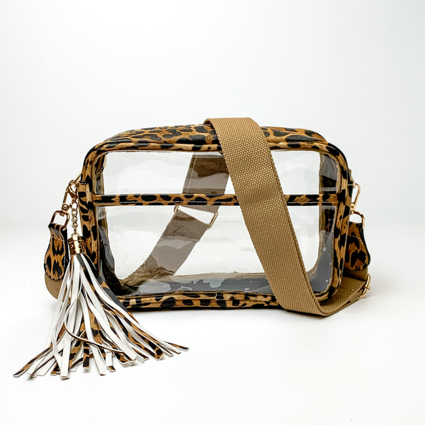 Pictured on a white background is a clear, rectangle purse with a leopard print outline. This purse includes gold accessories, a tan strap that is draped over the purse, and a fringe tassel that is white and leopard print. 