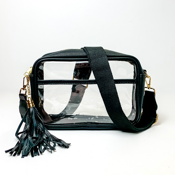 Pictured on a white background is a clear, rectangle purse with a black outline. This purse includes gold accessories, a black strap that is draped over the purse, and a black fringe tassel. 