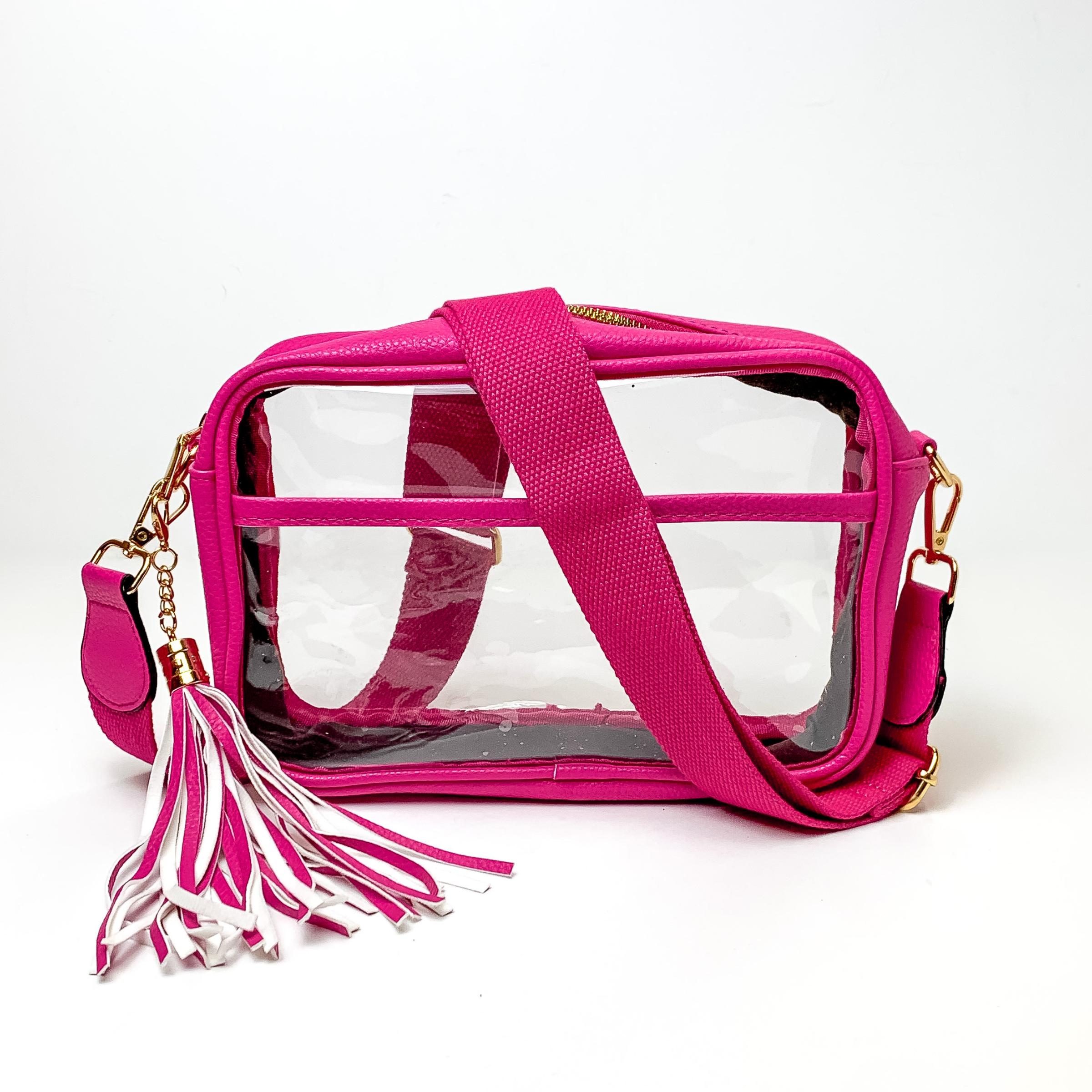 Pictured on a white background is a clear, rectangle purse with a fuchsia pink outline. This purse includes gold accessories, a fuchsia pink strap that is draped over the purse, and a pink and white fringe tassel. 
