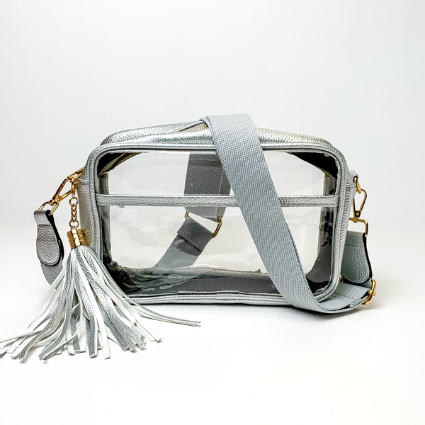 Pictured on a white background is a clear, rectangle purse with a silver outline. This purse includes gold accessories, a silver strap that is draped over the purse, and a silver and white fringe tassel. 