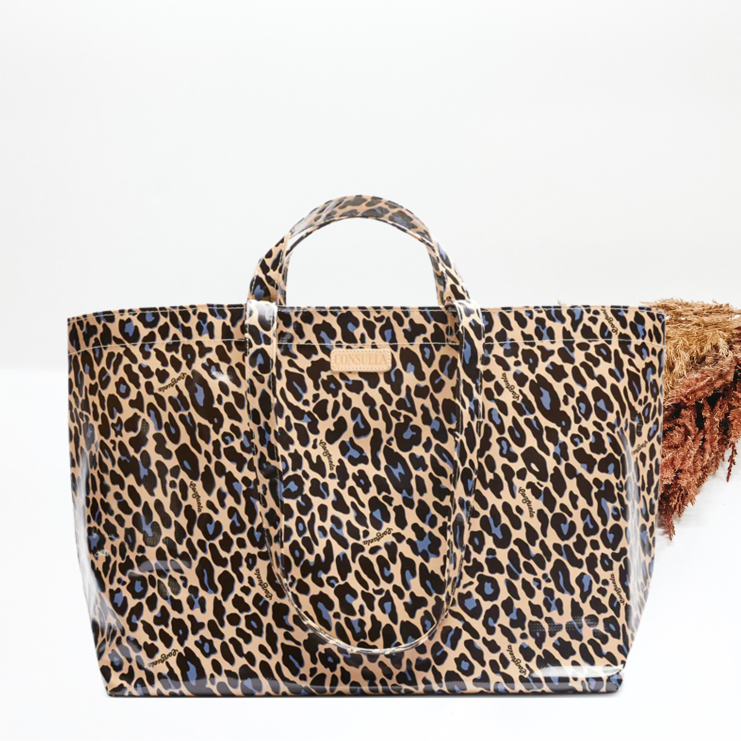 Large rectangle tote with short handles and long handles. This bag is tan in color with a black and blue leopard print. This tote is pictured in front of pompous grass on a white background.