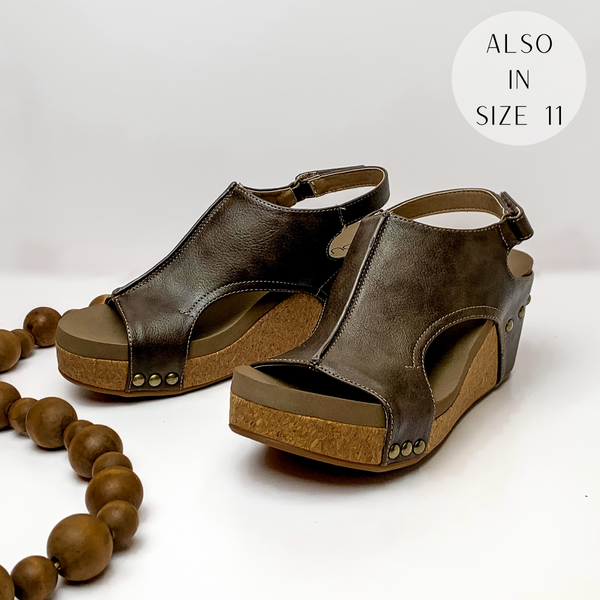 Cork wedges with a chocolate brown color upper with bronze studs connecting the upper to the wedge. These wedges also include a velco strap. These wedges are pictured on a white background with brown beads on the left side of the wedges.