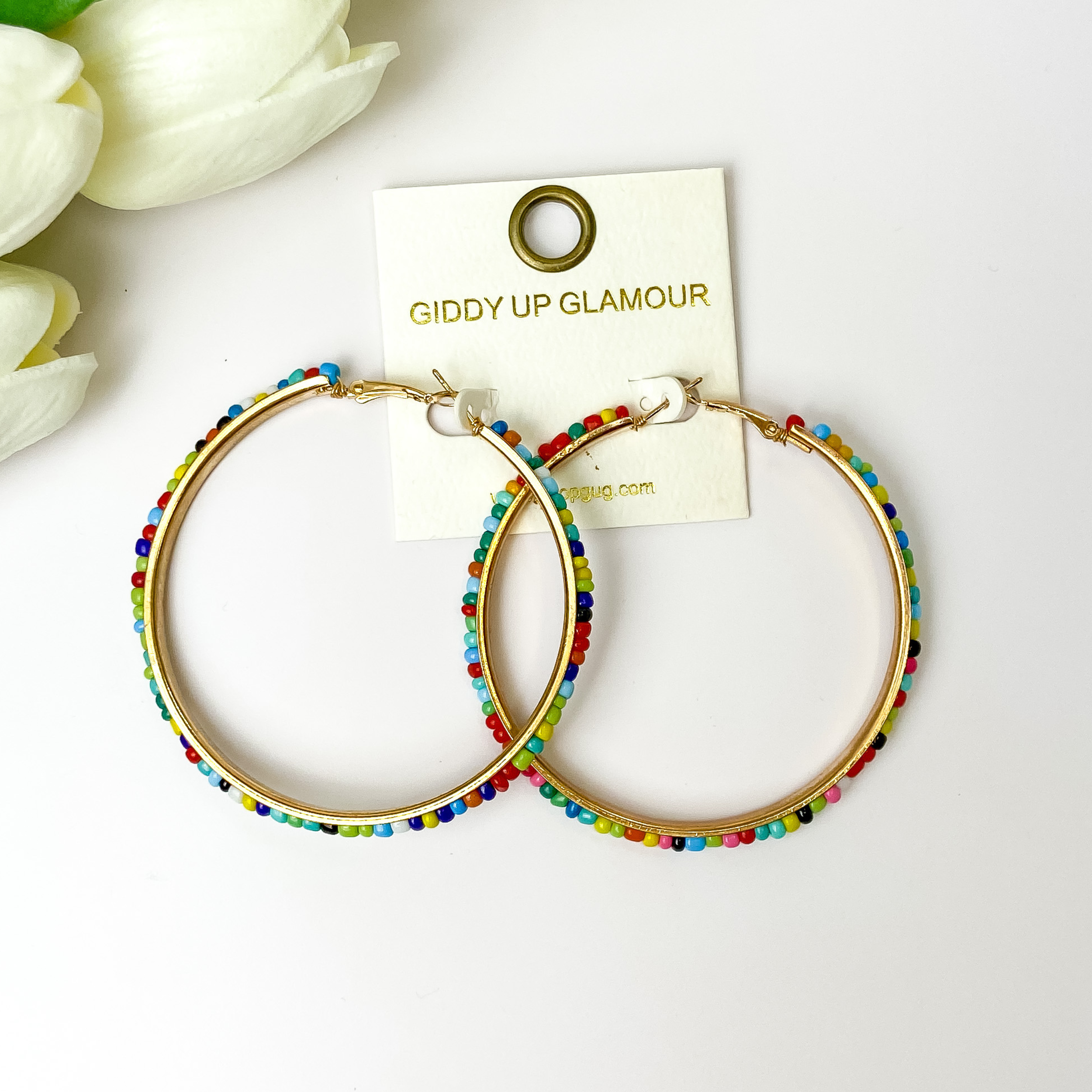 A pair of gold tone hoop earrings with multi color beads pictured on a white background with white flowers.