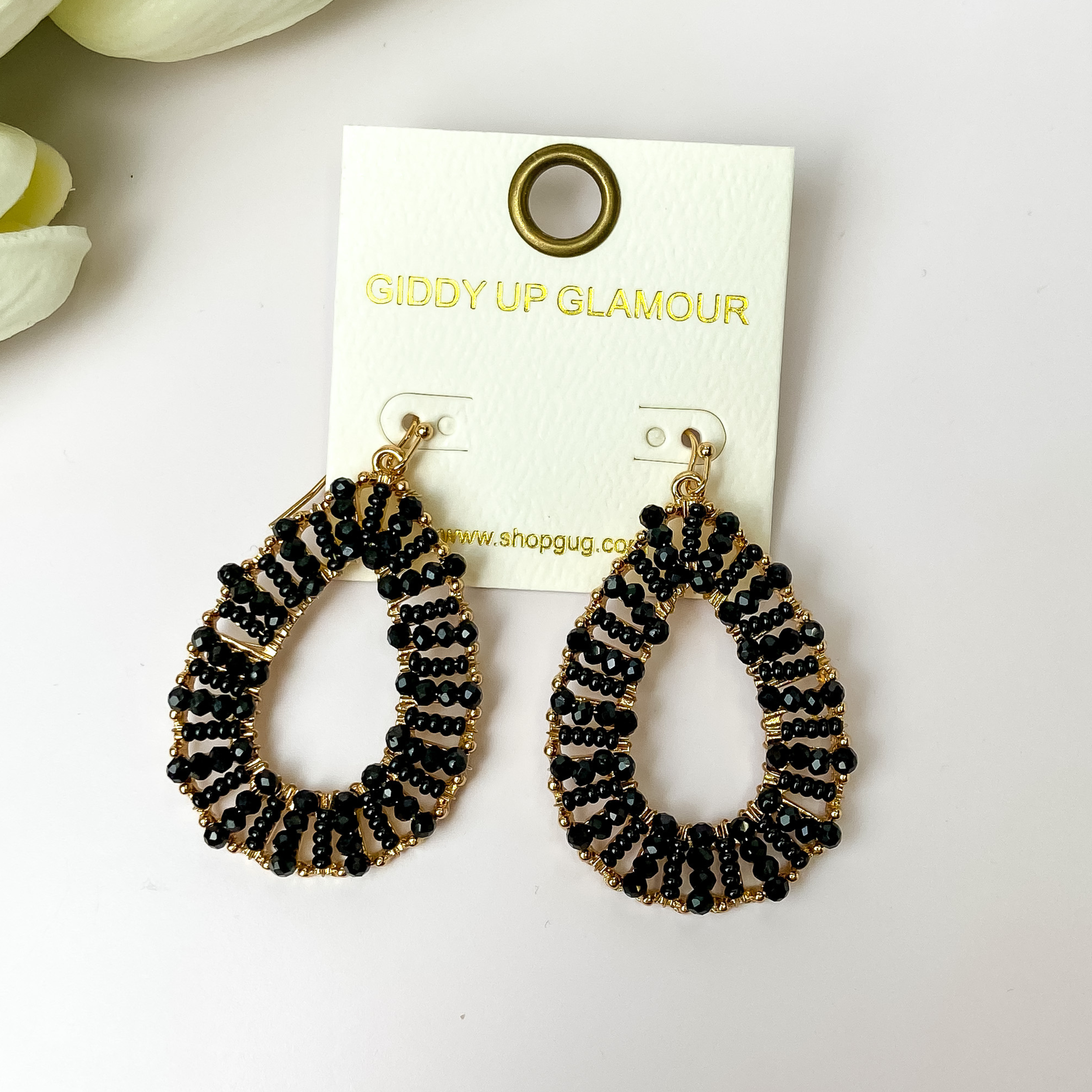 Gold undertone teardrop earrings with black beads pictured on a white background with white flowers.