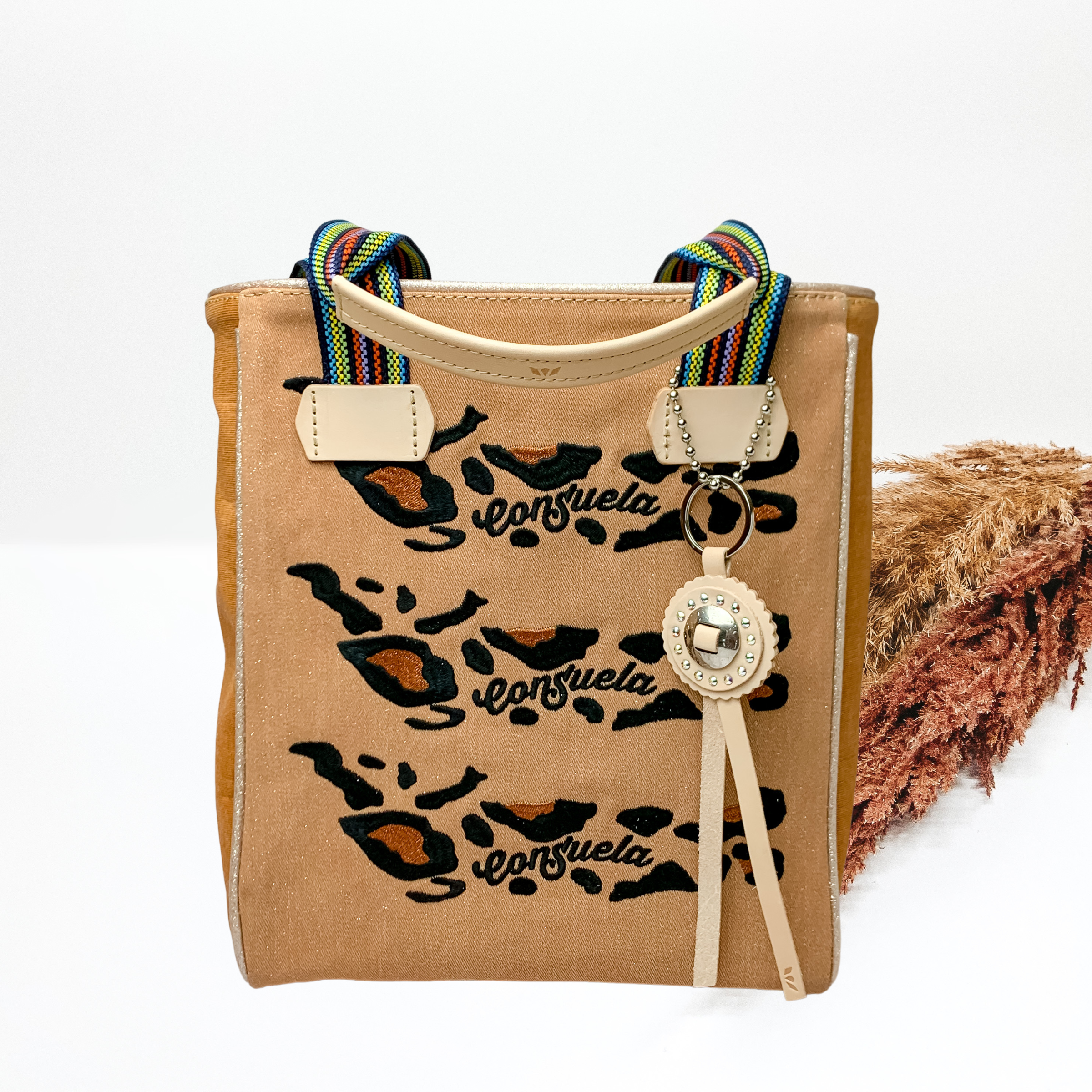 Rectangle tote bag with striped handles that have a light tan upper. This bag is a khaki color with a stitched brown and black leopard print and the word "CONSUELA" in black. This bag also includes a light tan circle charm with two light tan tassels. This bag is pictured in front of brown pompous grass on a white background. 