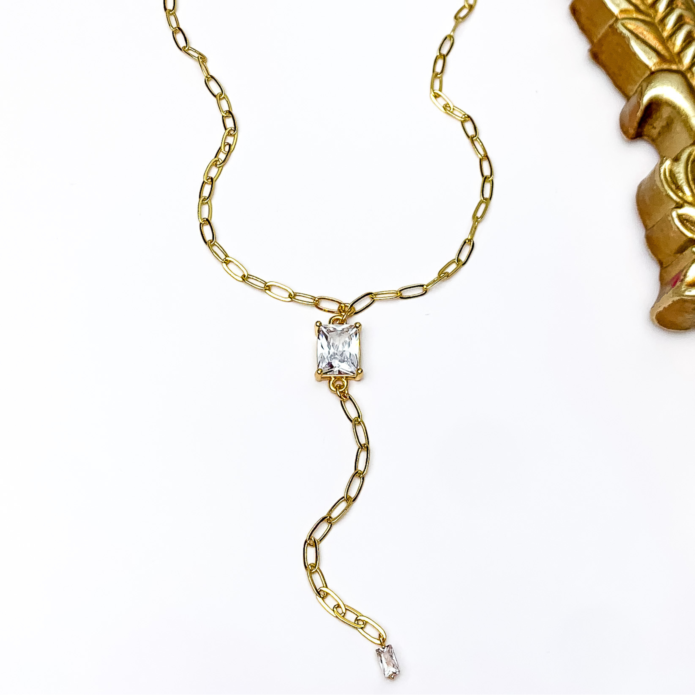 Pictured is a gold chain lariat neckalce with a rectabgle, clear crystal pendant and gold chain drop. This necklace is pictured on a white background with a gold mirror in the top right corner.  