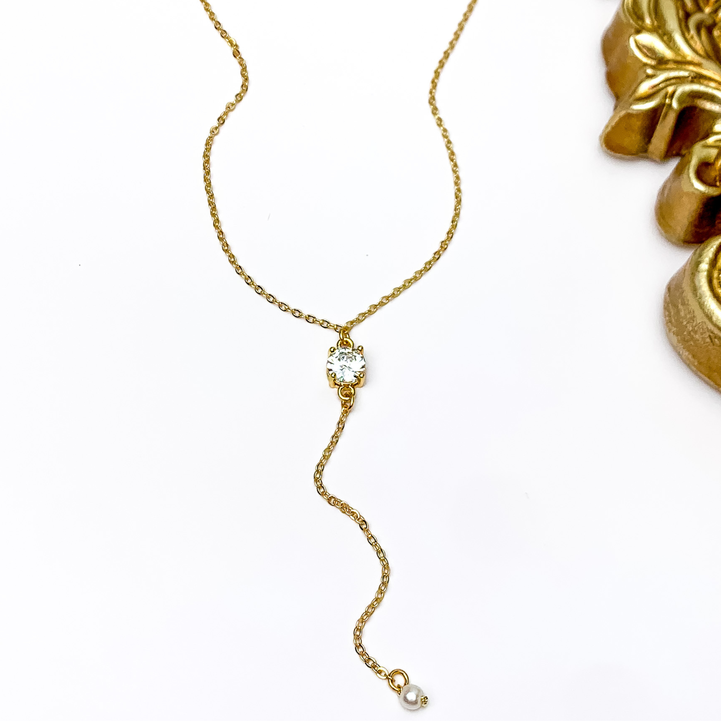 Pictured is a gold chain lariat neckalce with a rectabgle, clear crystal and white pearl pendant and gold chain drop. This necklace is pictured on a white background with a gold mirror in the top right corner.  