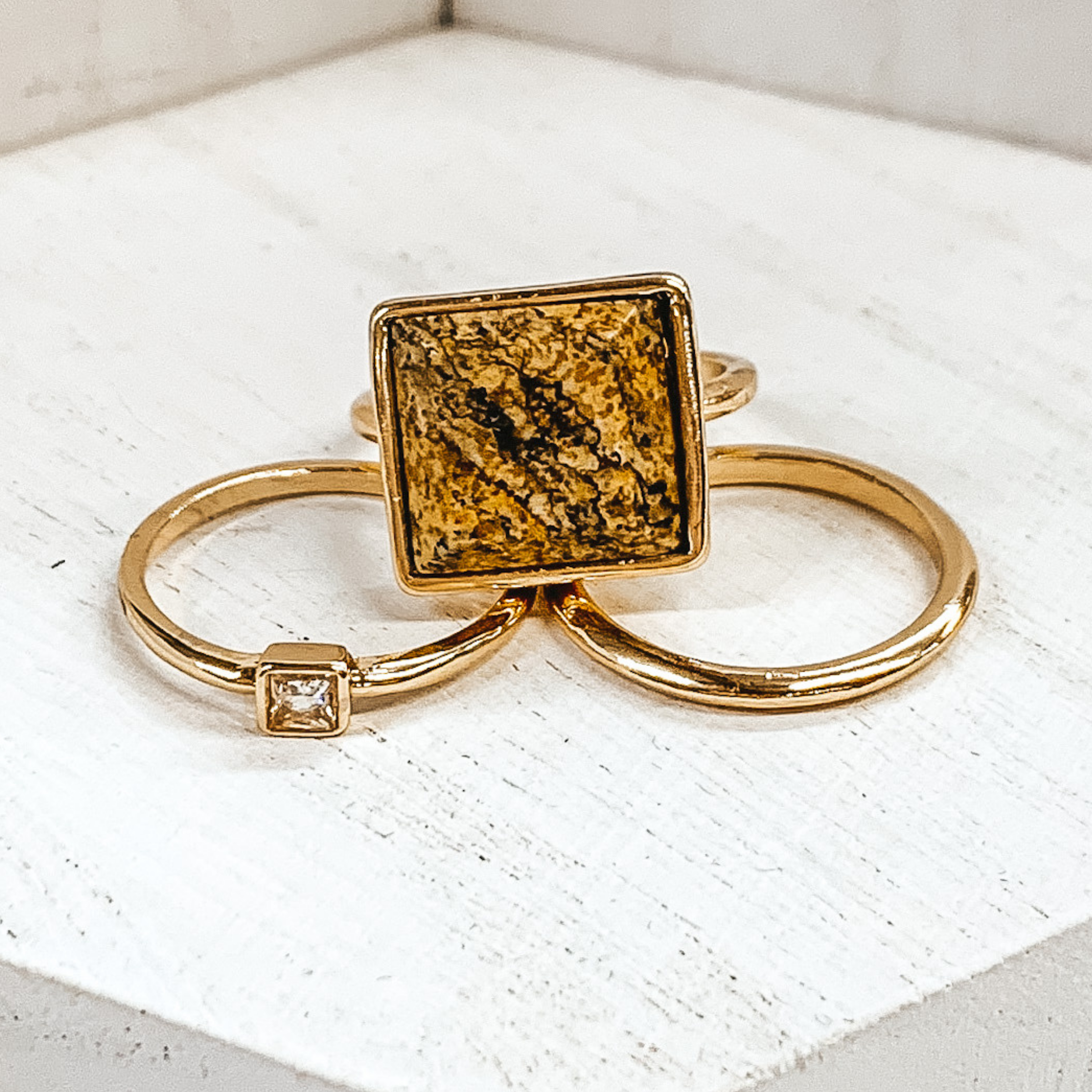 Set of three gold rings. One ring is a plain gold band. Another has a small square pendant with a center crystal. The third ring has a big, tan marbled pendant. These rings are pictured on a white background.  