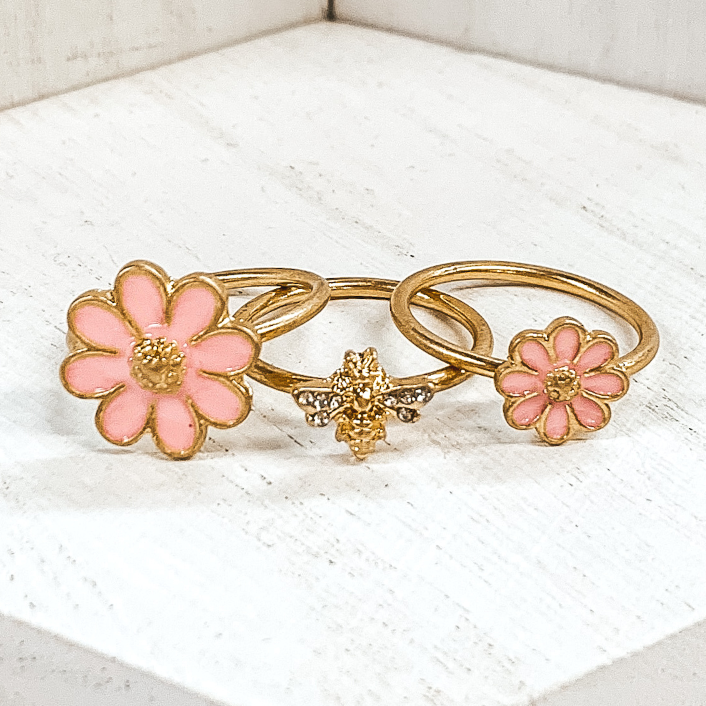 Set of three gold rings. Two rings have a pink flower pendant that is outlined in gold. One flower is bigger than the other. The last ring has a gold bee pendant with clear crystals on the wings. These rings are pictured on a white background. 