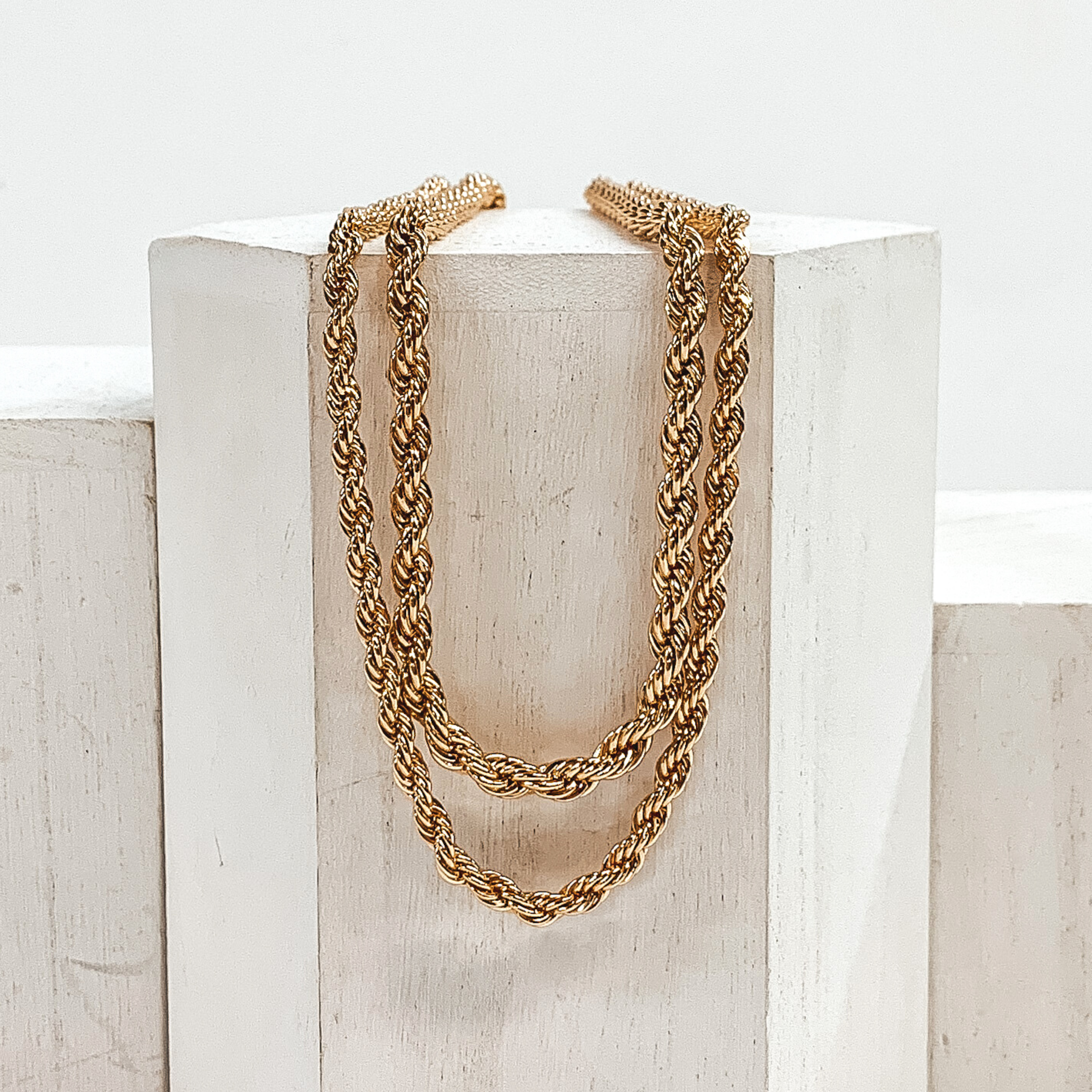 Two, gold, rope chain necklaces. This set includes a shorter and a longer necklace. This necklace is pictured laying on a white block on a white background.
