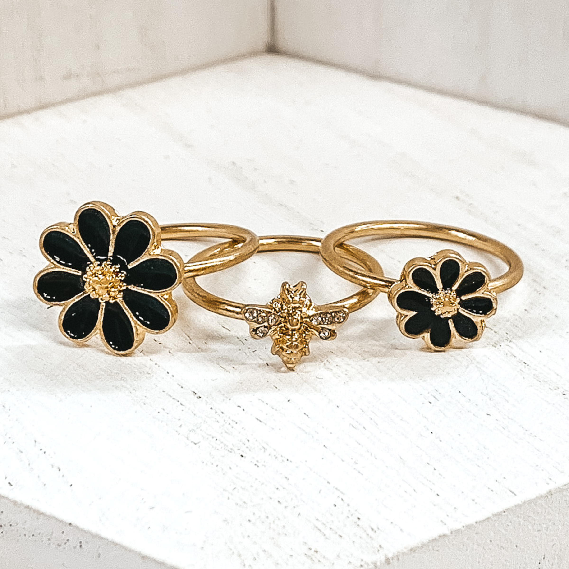Set of three gold rings. Two rings have a black flower pendant that is outlined in gold. One flower is bigger than the other. The last ring has a gold bee pendant with clear crystals on the wings. These rings are pictured on a white background. 