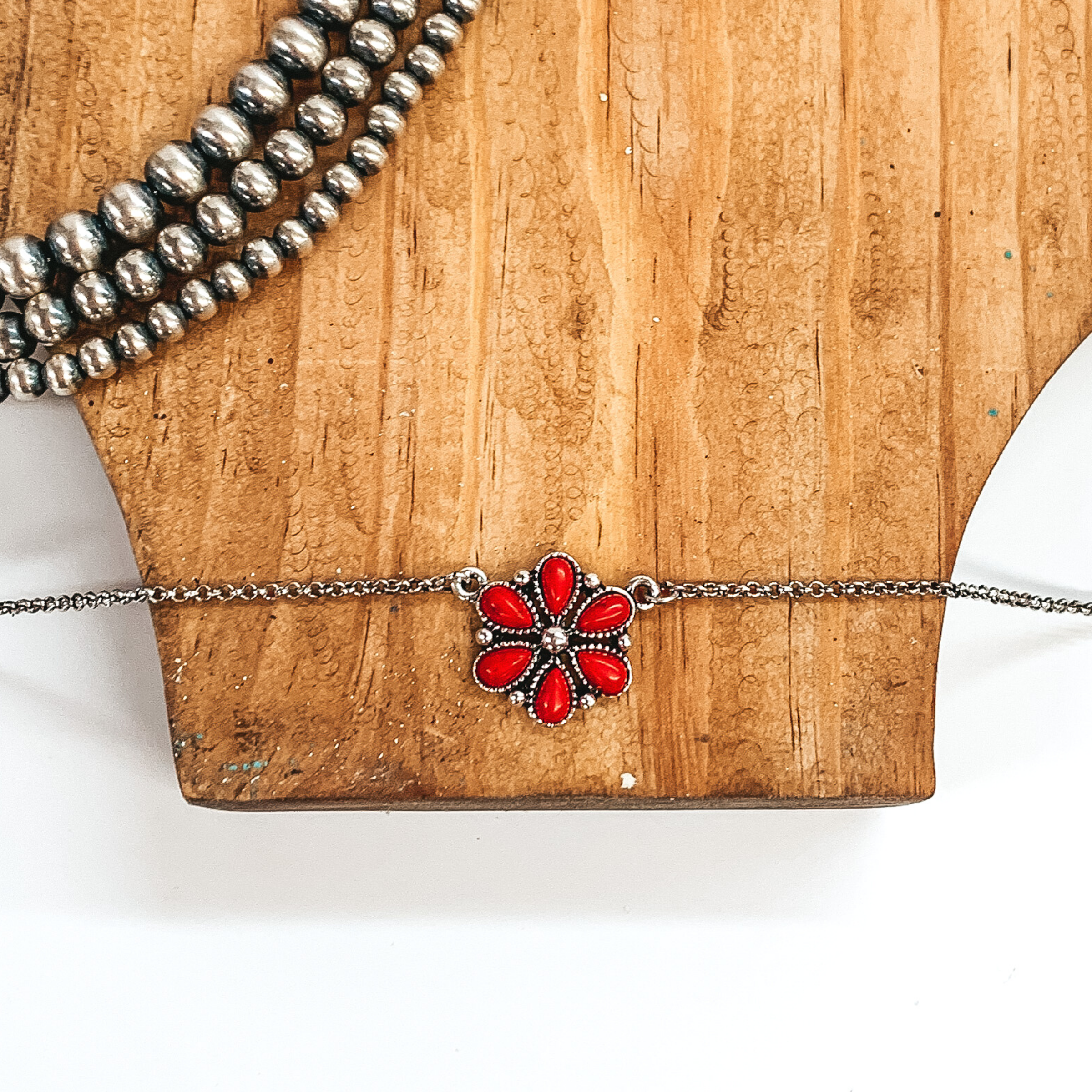 Silver chained necklace with a silver flower pendant that includes red stones. This necklace is pictured laying on a brown necklace holder with silver pearls in the top left corner as decoration