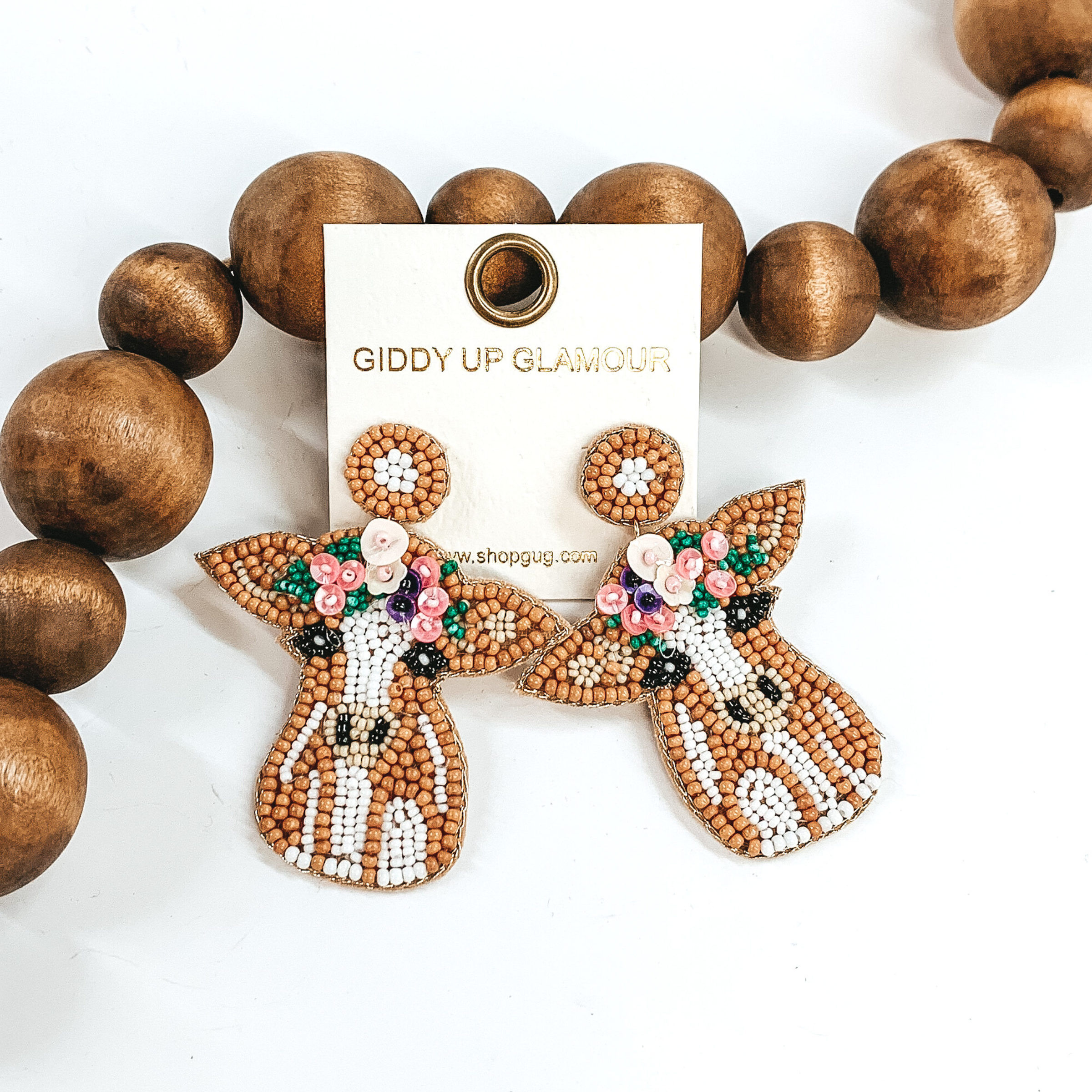 Beaded cow dangle earrings with a post back. The cow is a tan and white detailed cow. There is also beaded flower detailing on the cow head. These earrings are pictured on a white background with brown beads behind it. 