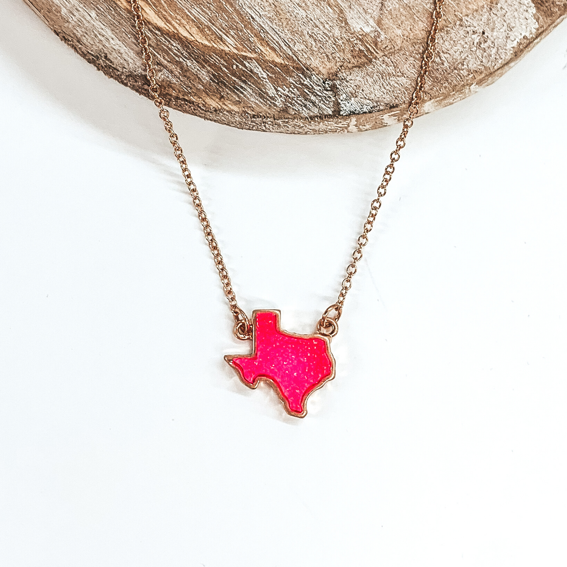 Tiny gold chained necklace includes a neon pink, druzy texas pendant that is outlined in gold. This necklace is pictured on a white background with a tan piece of wood at the top of the picture.
