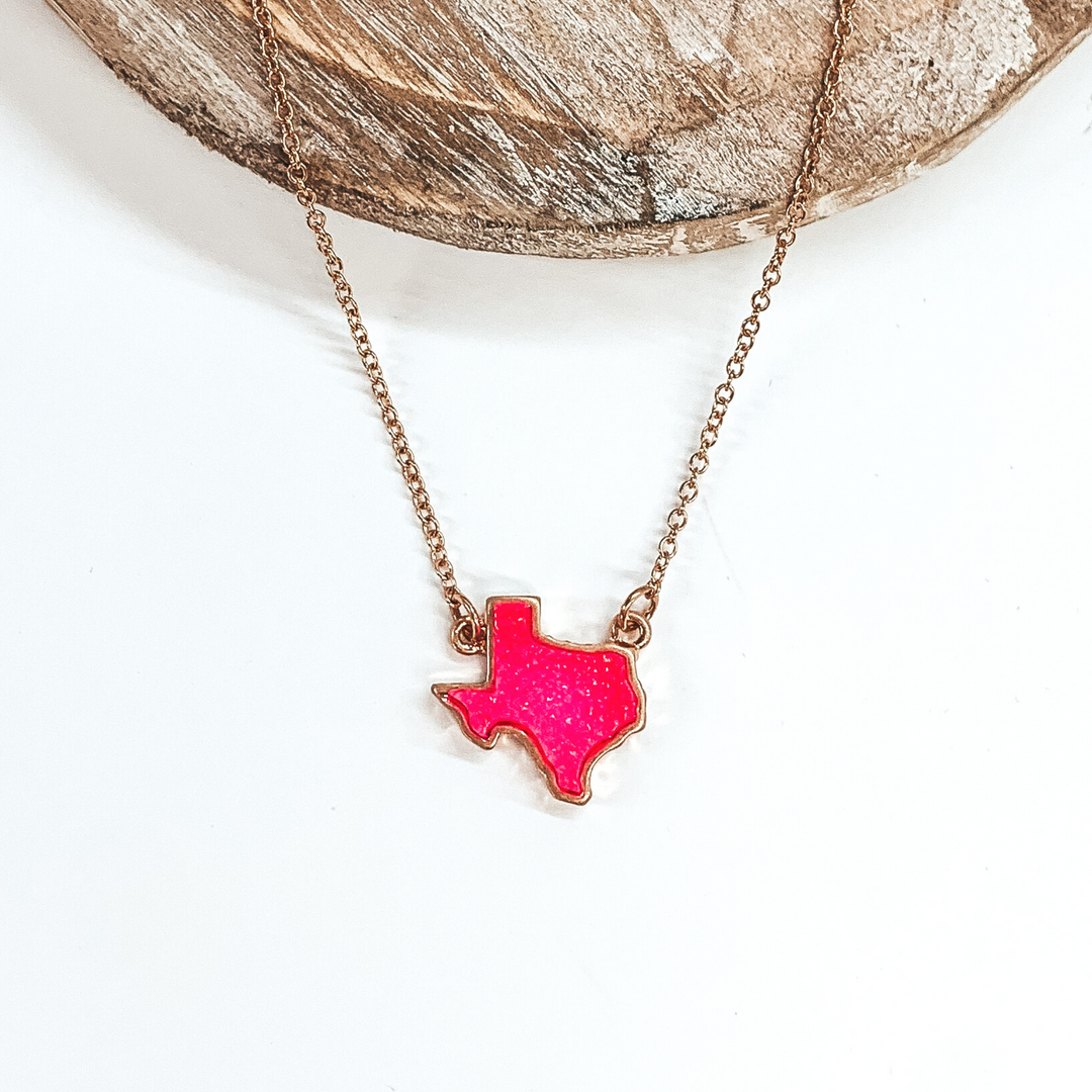 Tiny gold chained necklace includes a neon pink, druzy texas pendant that is outlined in gold. This necklace is pictured on a white background with a tan piece of wood at the top of the picture.