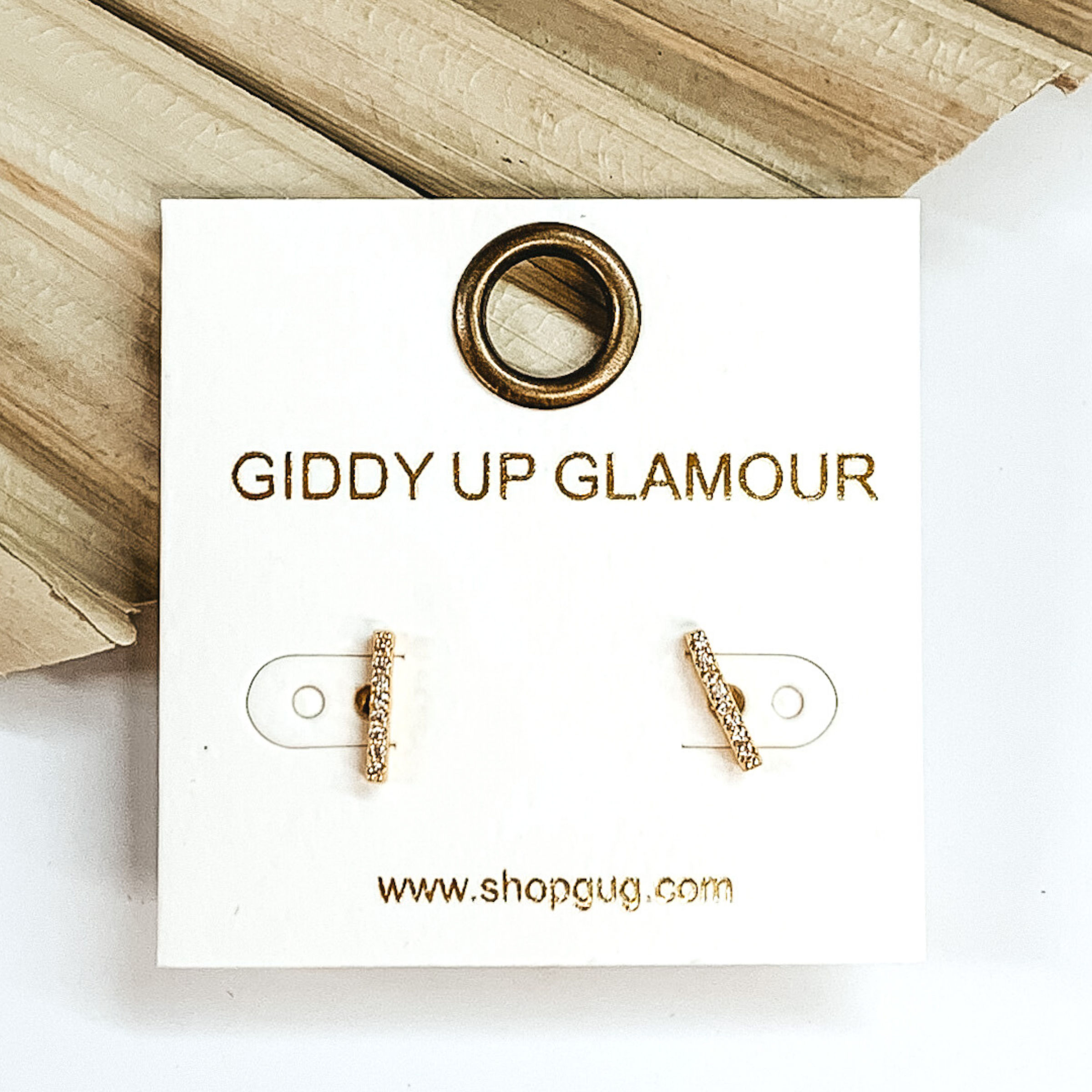 Gold and clear crystals mini bar stud earrings. These earrings are pictured laying on a green palm leaf on a white background.