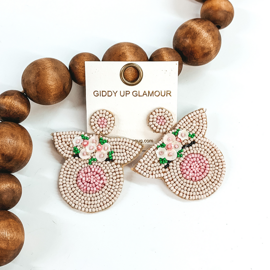 These are beaded earrings are in the shape of a pig head with pale pink and pink detailing. There is also beaded flower detailing on the pig head. These earrings are pictured on a white background with brown beads behind it.
