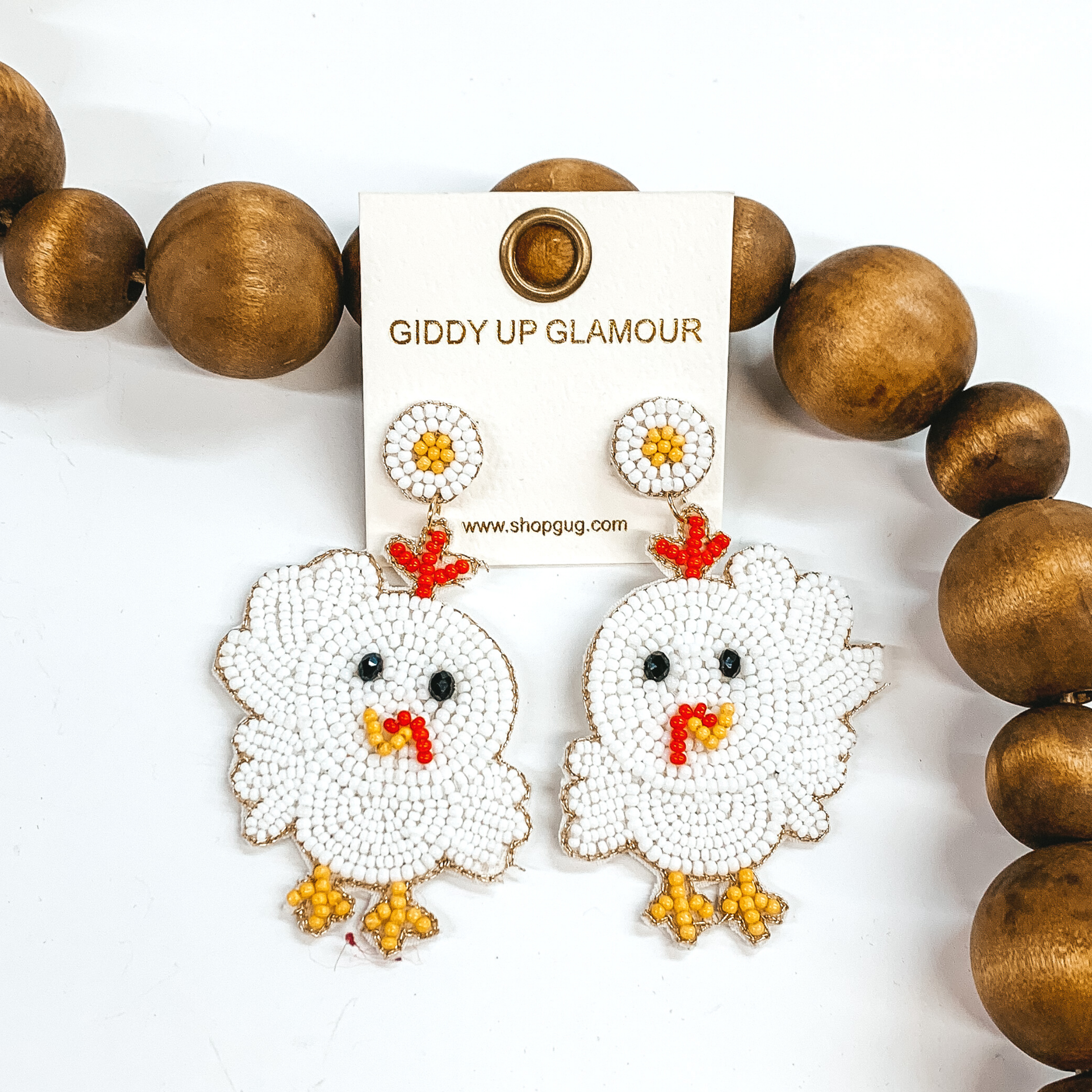 These are beaded chick shaped dangle earrings with a post back. These earrings are white with red and yellow detailing. These earrings are pictured on a white background with brown beads behind it.