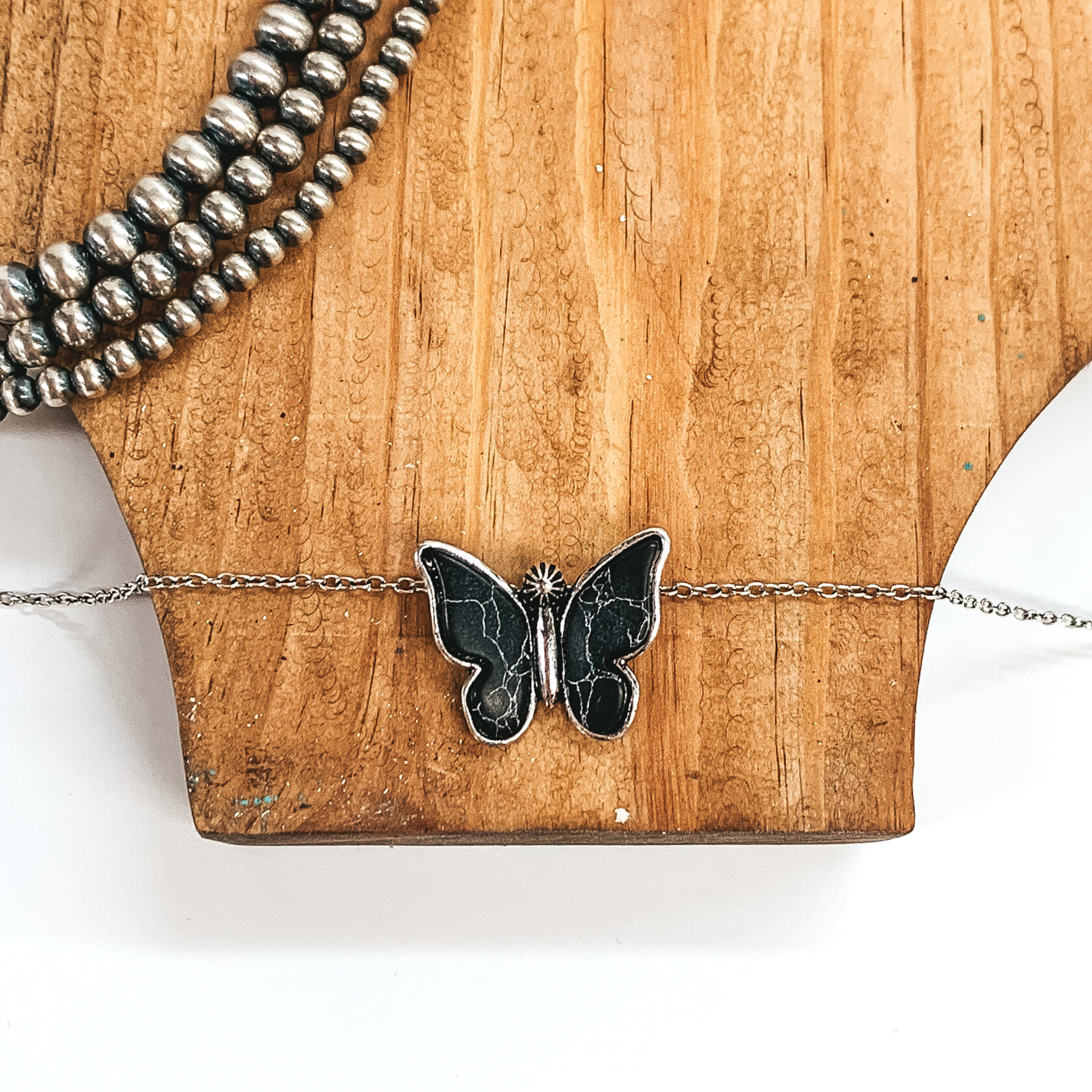 Silver chained necklace with a silver butterfly pendant that includes black stones. This necklace is pictured laying on a brown necklace holder with silver pearls in the top left corner as decoration. 