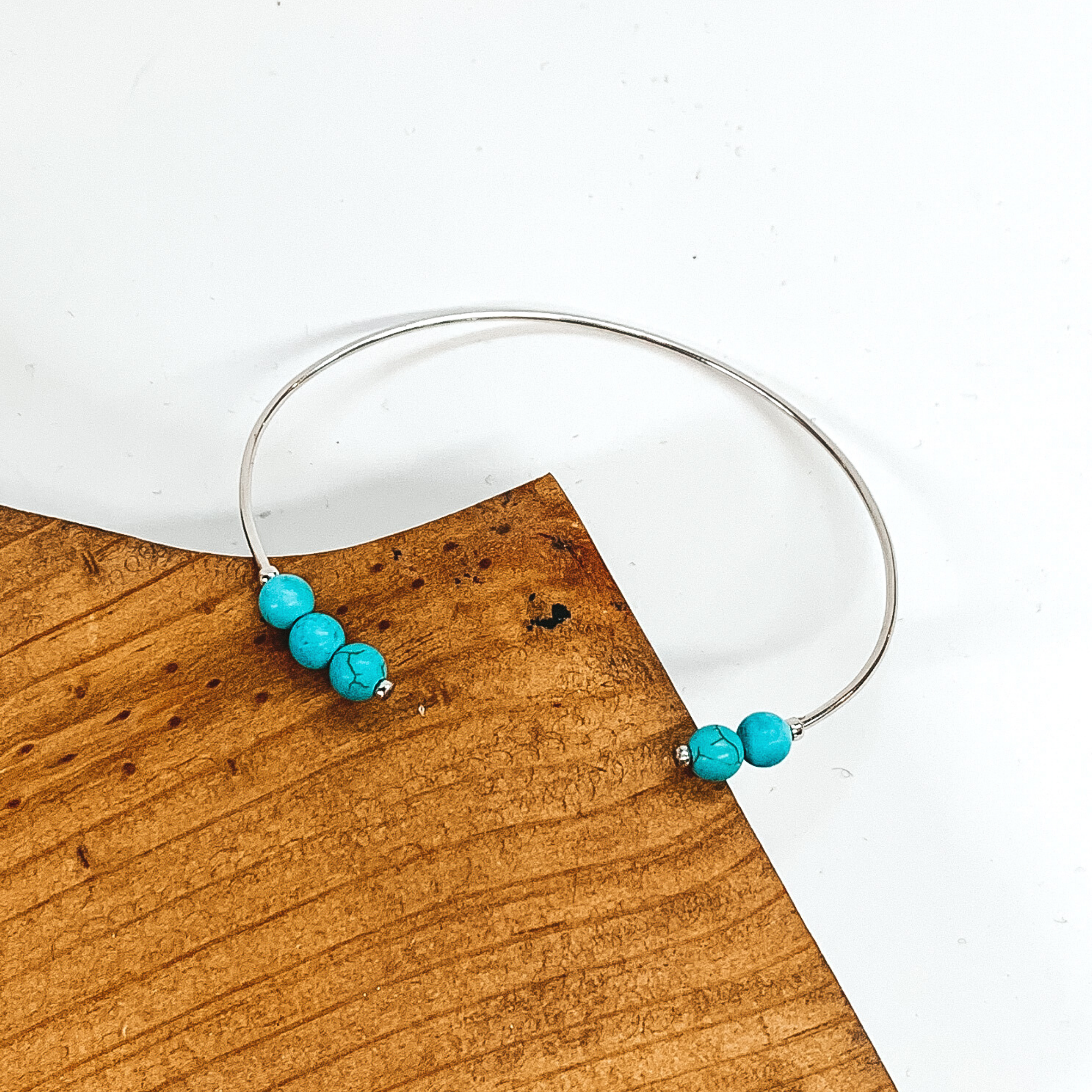 Silver, thin wire bangle bracelet with beaded turquoise ends. This bracelet is pictured laying partially on a brown block on a white background. 