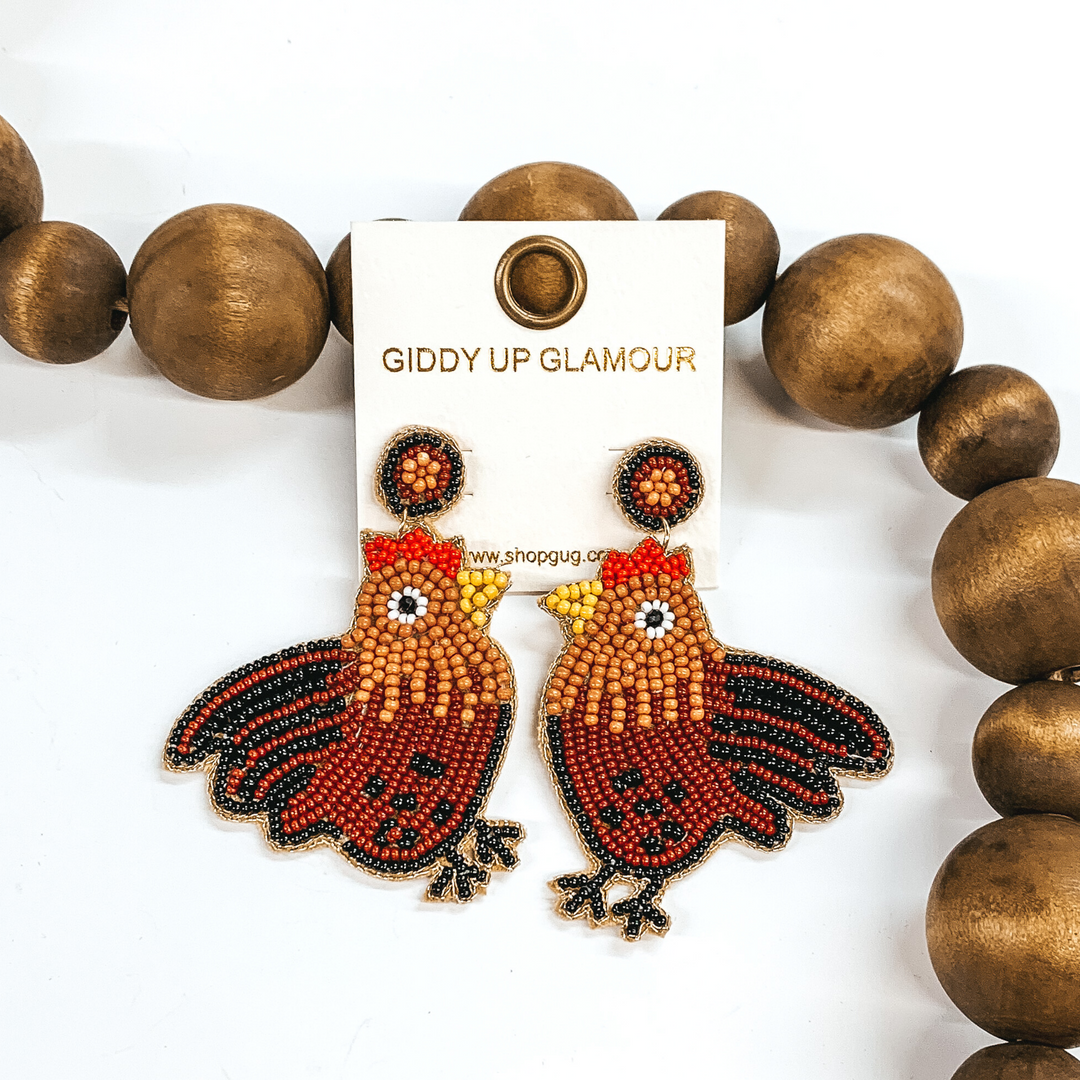 These earrings are post back dangle earrings in the shape of a rooster. The rooster has brown, black, and tan detailing. These earrings are pictured on a white background with brown beads behind it.