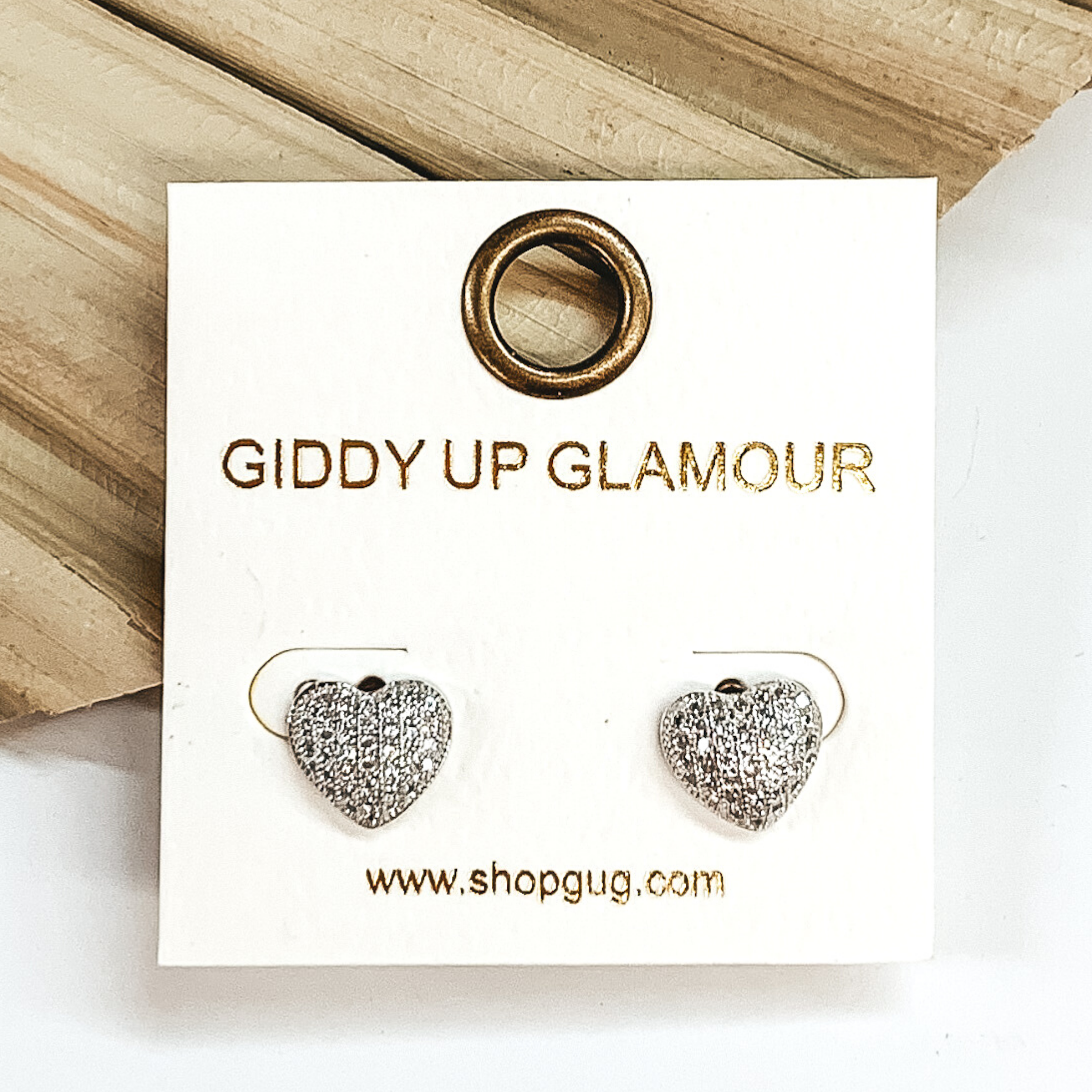 Clear crystal heart stud earrings in silver. These earrings are pictured laying on a green palm leaf on a white background.