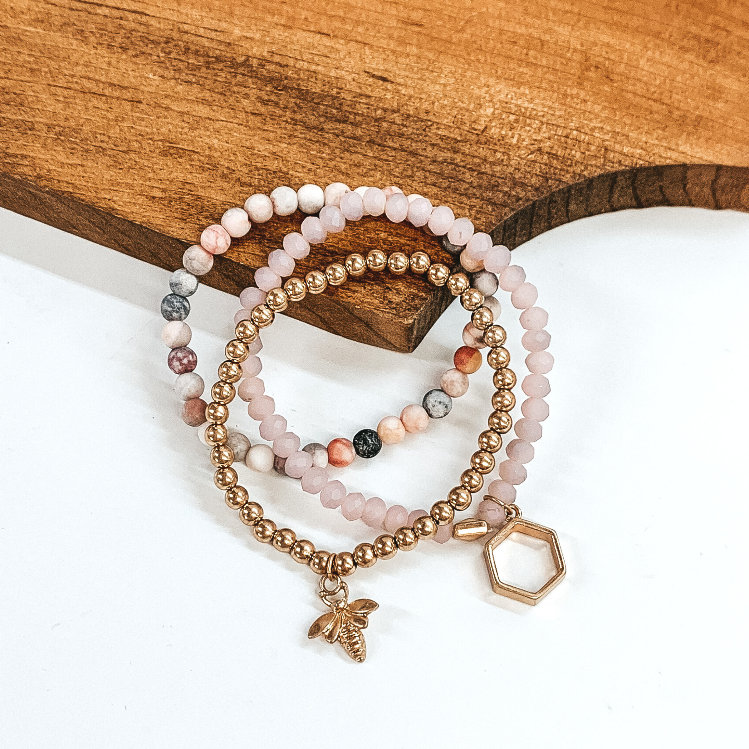 Set of three beaded bracelets with a gold bee pendant and a gold hexagon pendant. This set includes a light blush bracelet, a pink marbled beaded bracelet, and a gold beaded bracelet. These bracelets are pictured leaning on a brown block on a white background. 