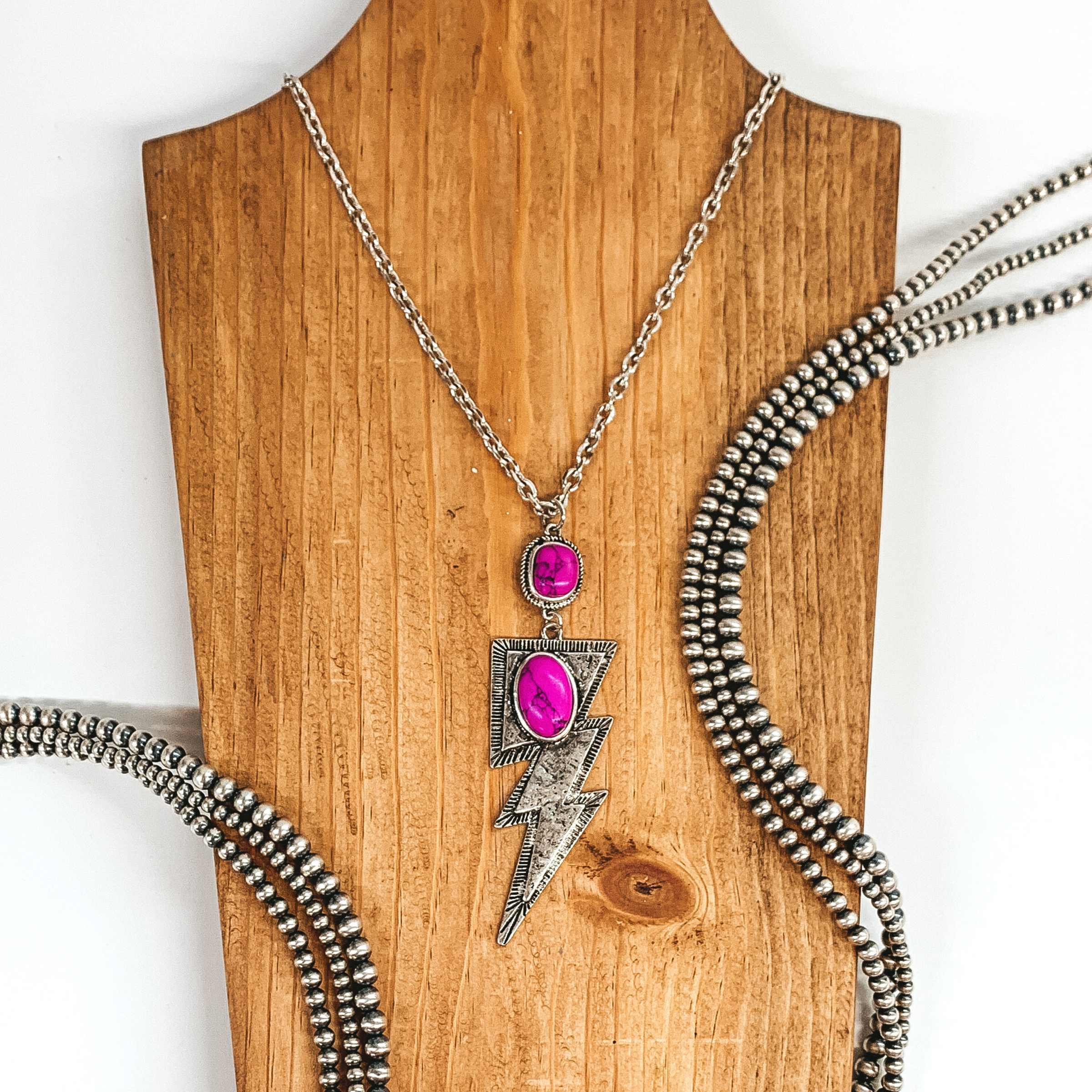 Silver chained necklace with hanging square pendant with fuchsia stone and hanging silver lightning bolt pendant with oval fuchsia colored stone. This necklace is pictured laying on a tan necklace holder on a white background. This picture includes silver beads as decoration.