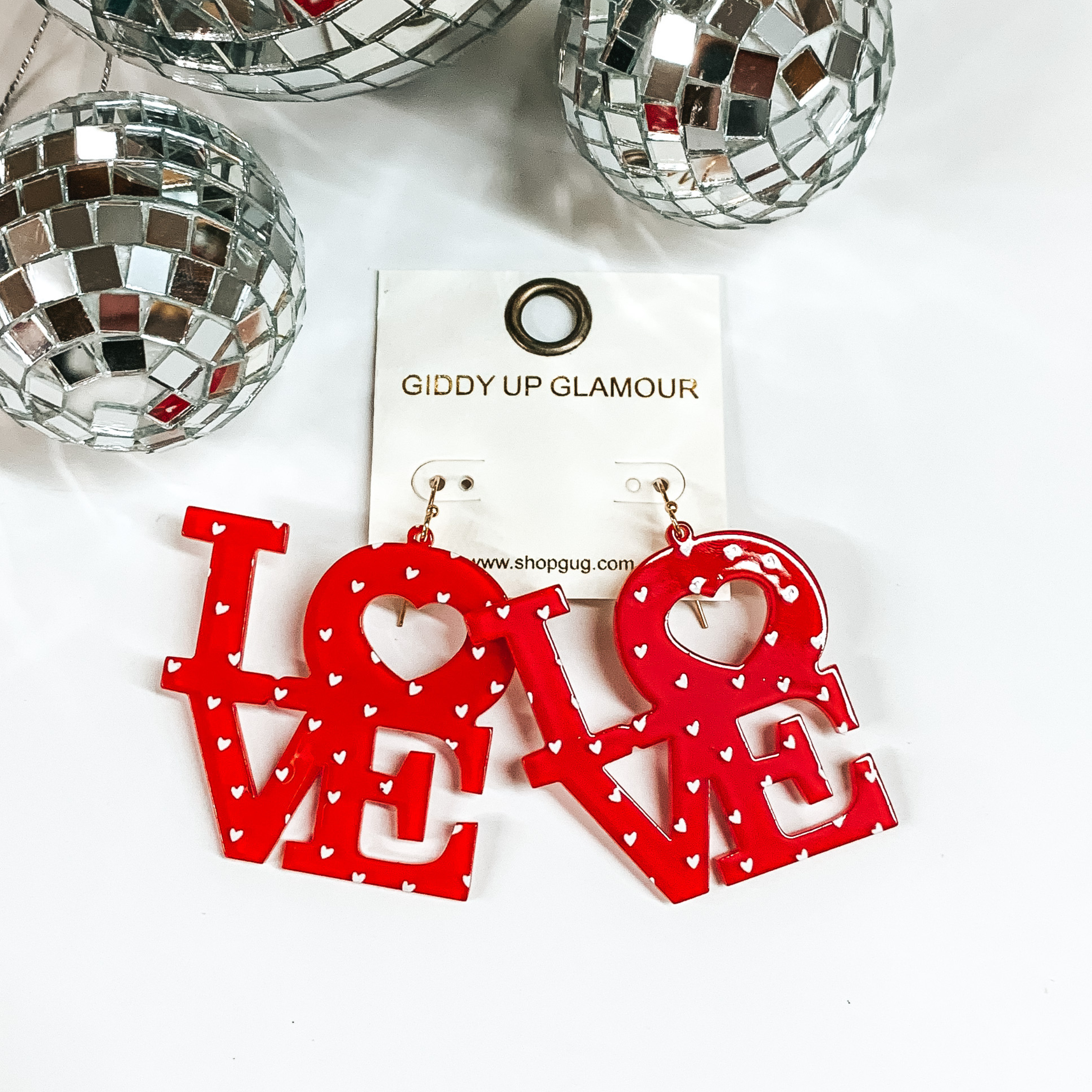 Gold fish hook with red block letters that spell out "LOVE". These earrings also include small, white hearts throughout the letters. These earrings are pictured on a white background with disco balls in the top left corner. 