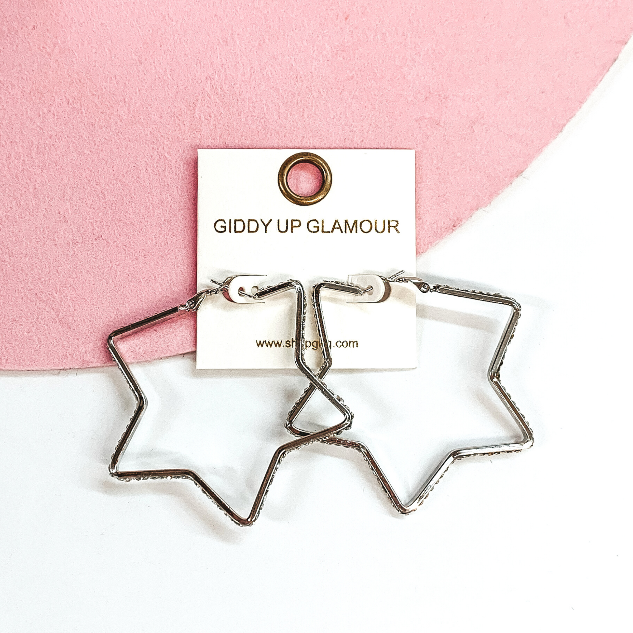 Silver star shaped hoop earrings with clear crystals outlining the outside of the hoops. These earrings are pictured on a white and pink background. 