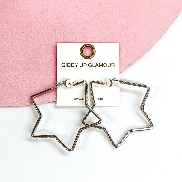 Silver star shaped hoop earrings with clear crystals outlining the outside of the hoops. These earrings are pictured on a white and pink background. 