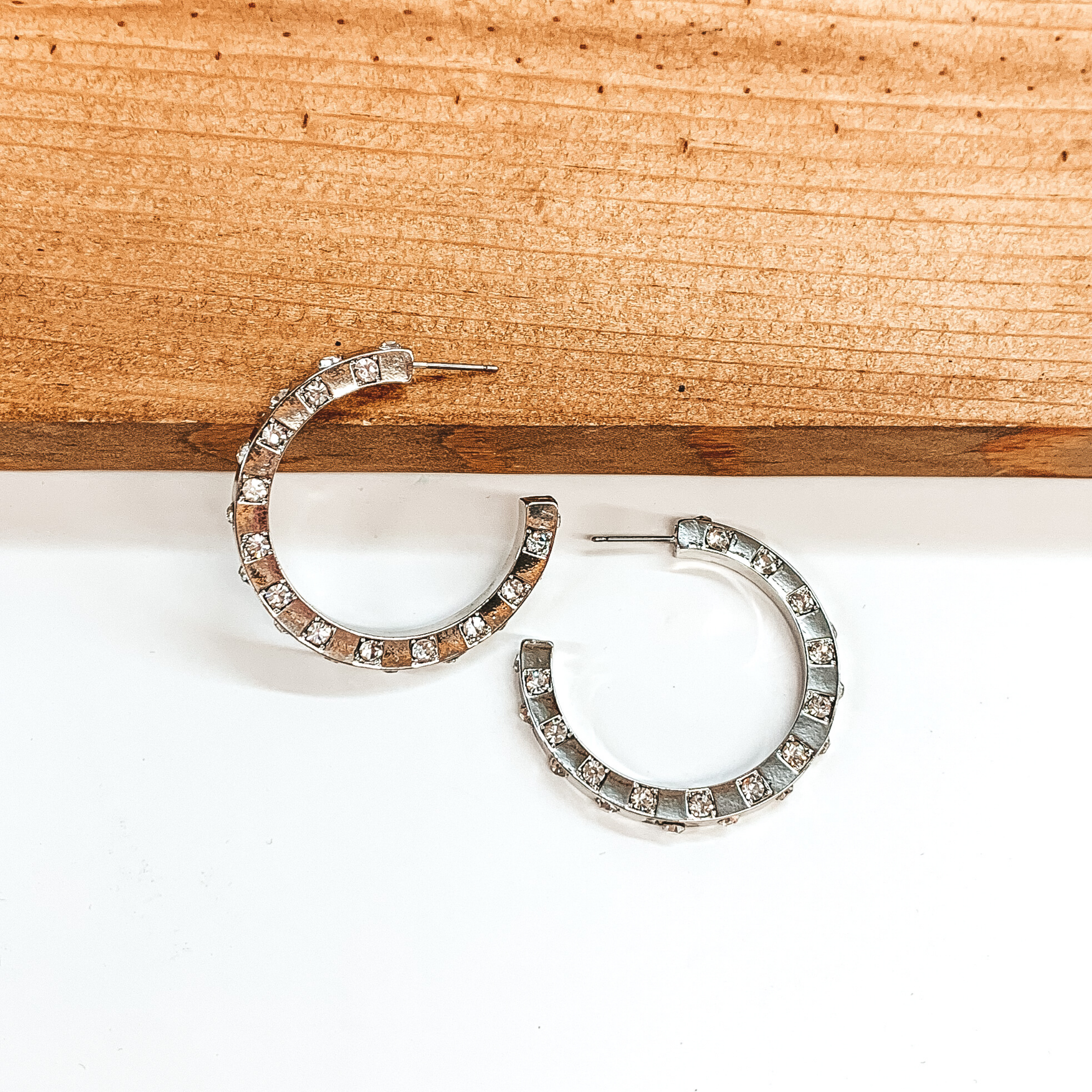 Silver hoop earrings with spaced out clear crystals. One earrings is pictured laying on a brown block while the other earrings is pictured laying flat on the white background. 