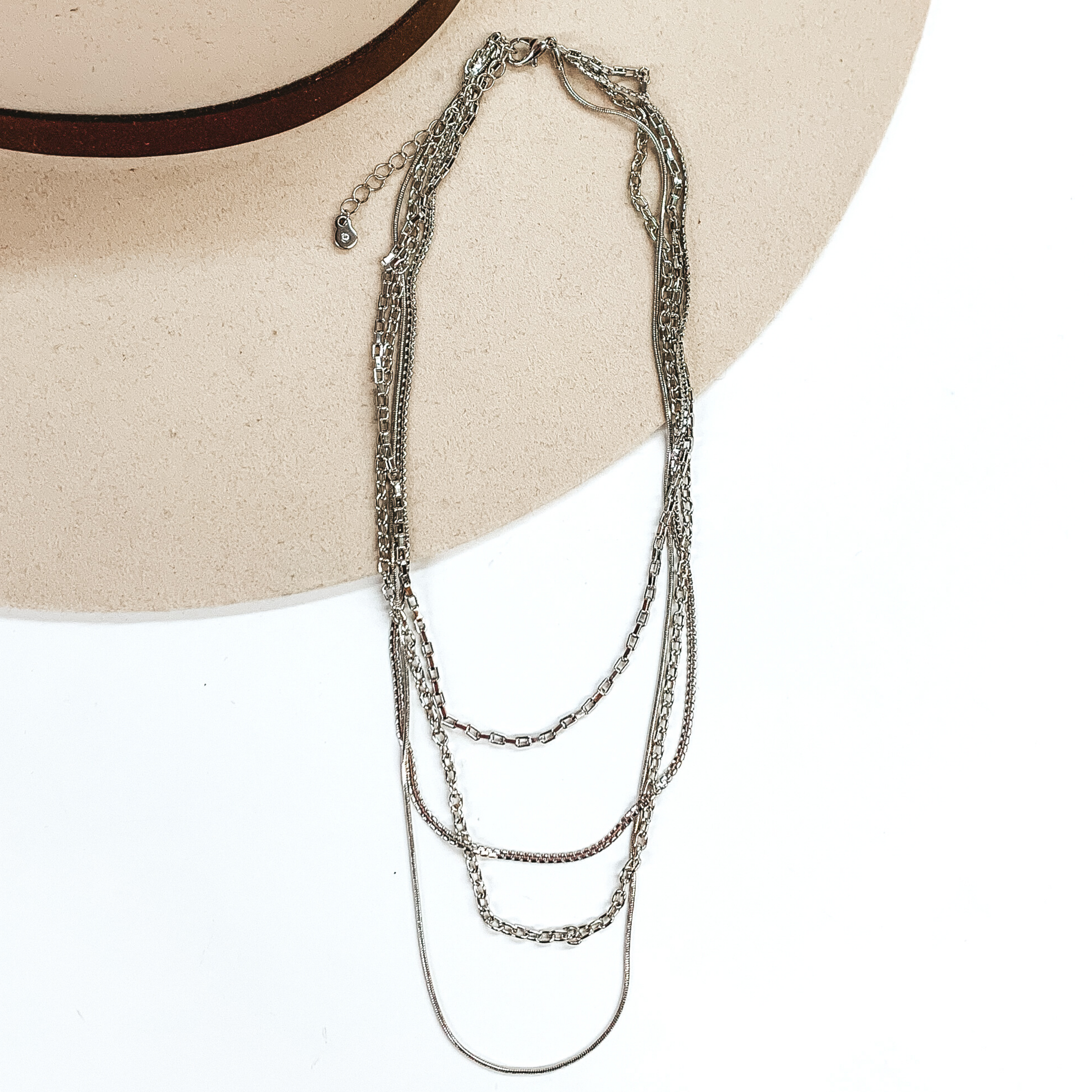 This is a silver, four stranded necklace that has different types of chains at different lengths. These necklaces are pictured partially laying on top of a beige hat brim on a white background.