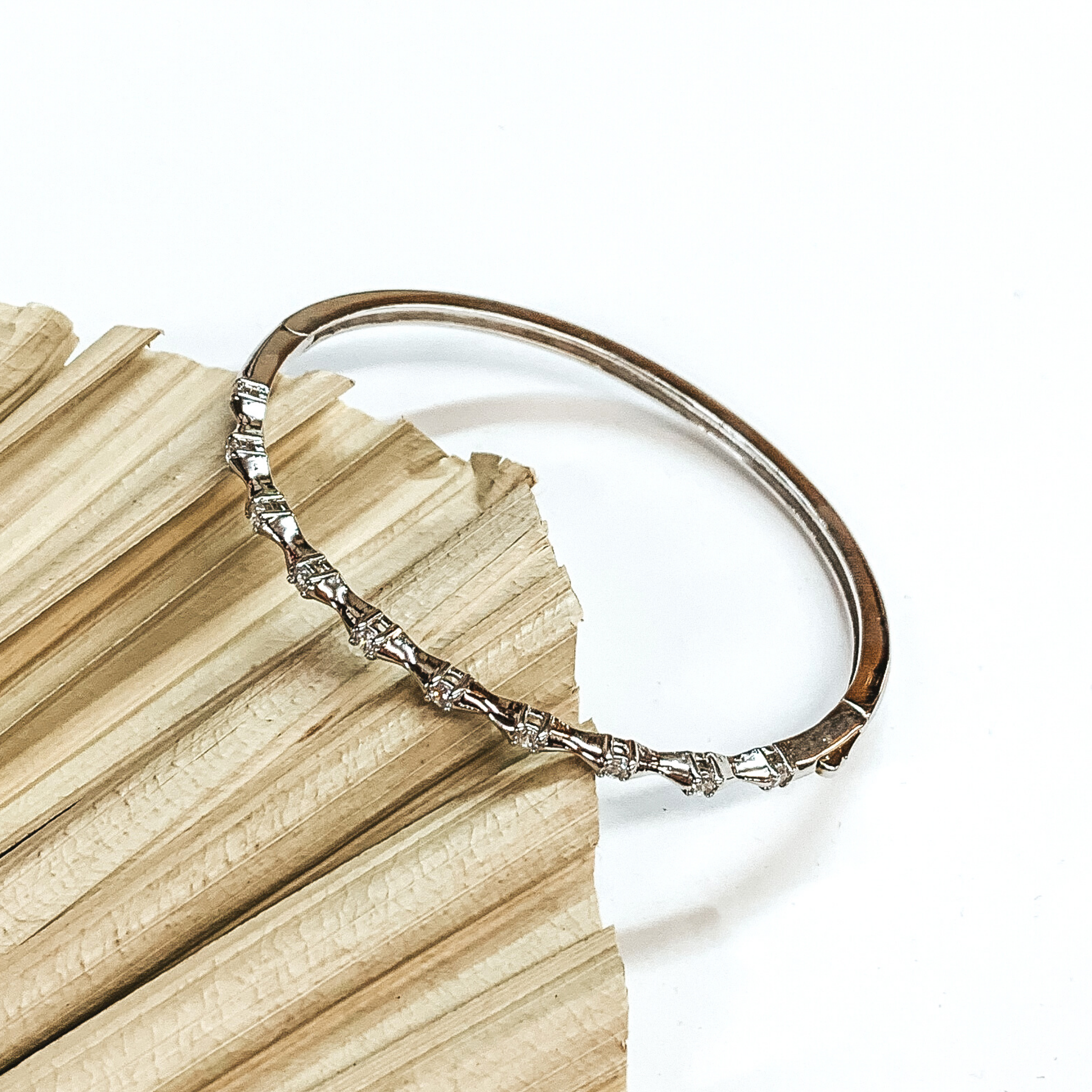 This is a thin silver bangle bracelet with clear crystal spacers. This bracelet is pictured laying on a green palm leaf on a white background.