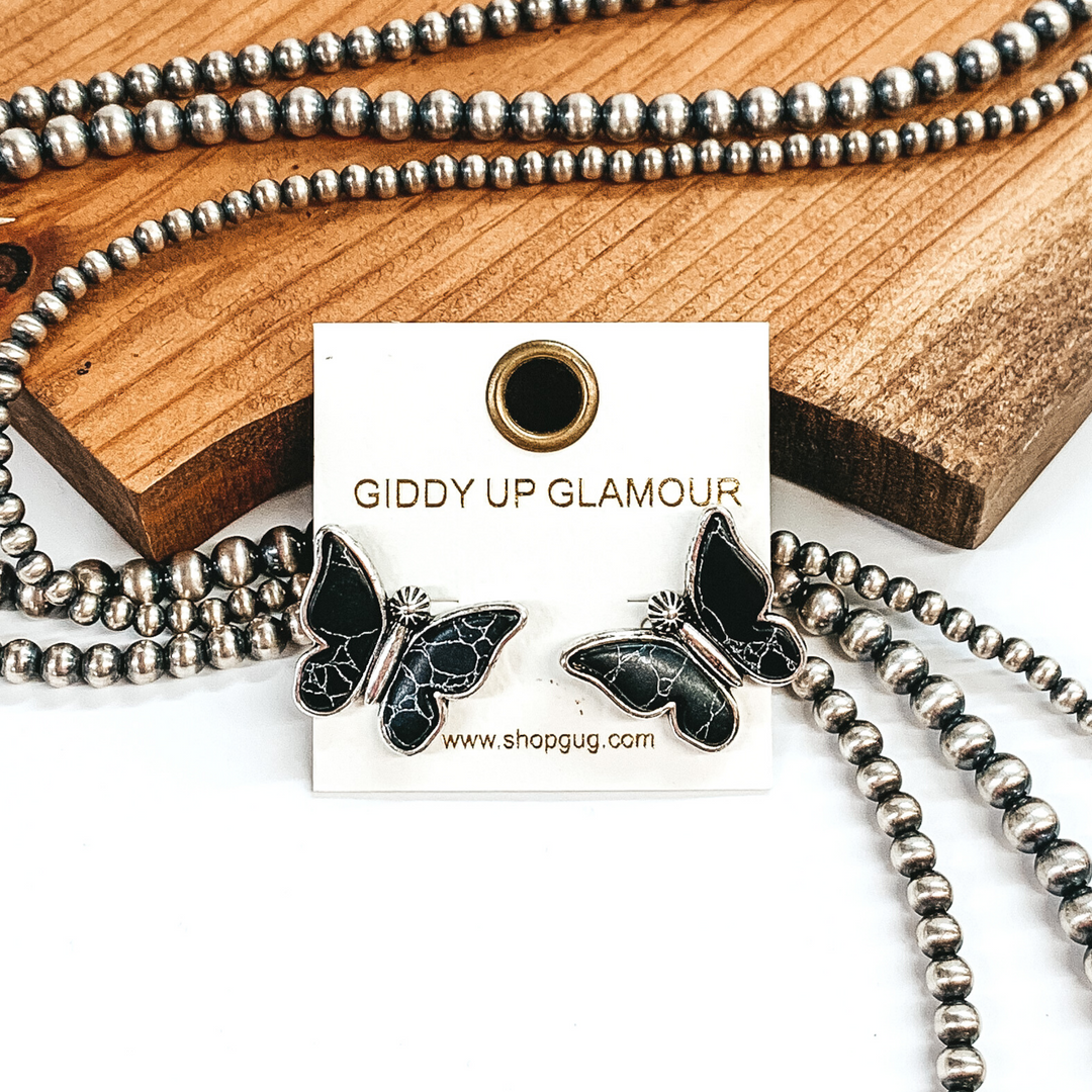 Silver post stud earrings shaped like a butterfly with black stones for wings. These earrings are pictured in front of brown wood with some silver pearls laying behind the earrings. 